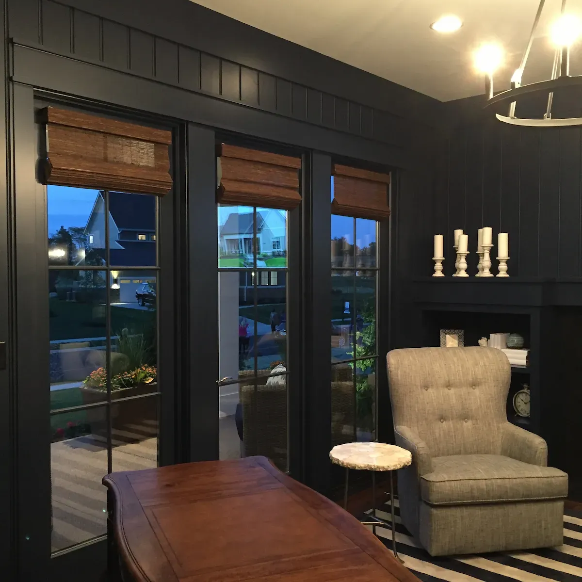 The image shows an interior view of a house at dusk, looking out through three tall windows with dark frames. The upper portion of each window is fitted with rich, brown woven wood shades, partially drawn. Outside the windows, a residential neighborhood with houses and green lawns is visible under the twilight sky. Inside, the room has dark paneled walls. To the right, a comfortable-looking upholstered armchair with a small, round side table sits next to a built-in shelf unit decorated with candles and other items. A large, dark wooden desk or table is in the foreground on the left. The room is illuminated by overhead lights and possibly a chandelier, casting a warm glow.