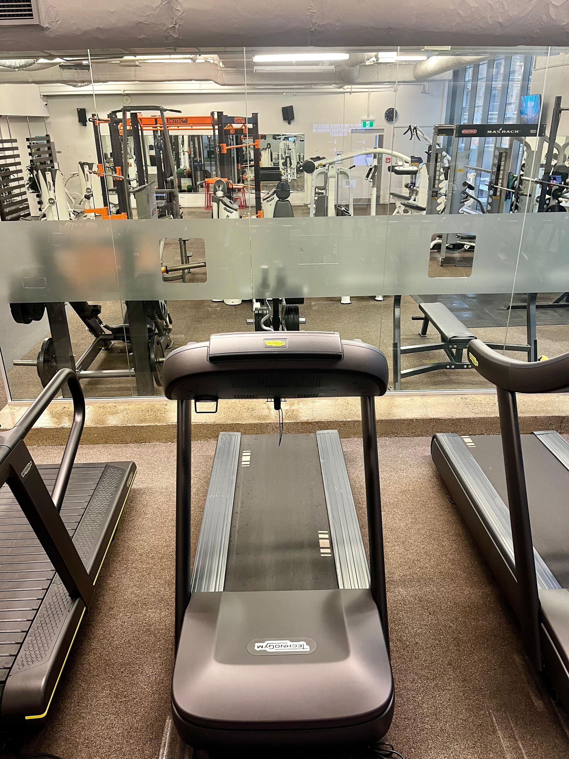 Three treadmills in a gym, with other workout equipment visible through a glass wall.