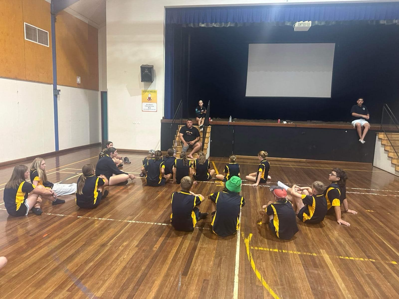 Children in School Uniforms Seated on a Wooden Floor, Listening to an Adult Speaker — Dinawans Connection in Cobar, NSW