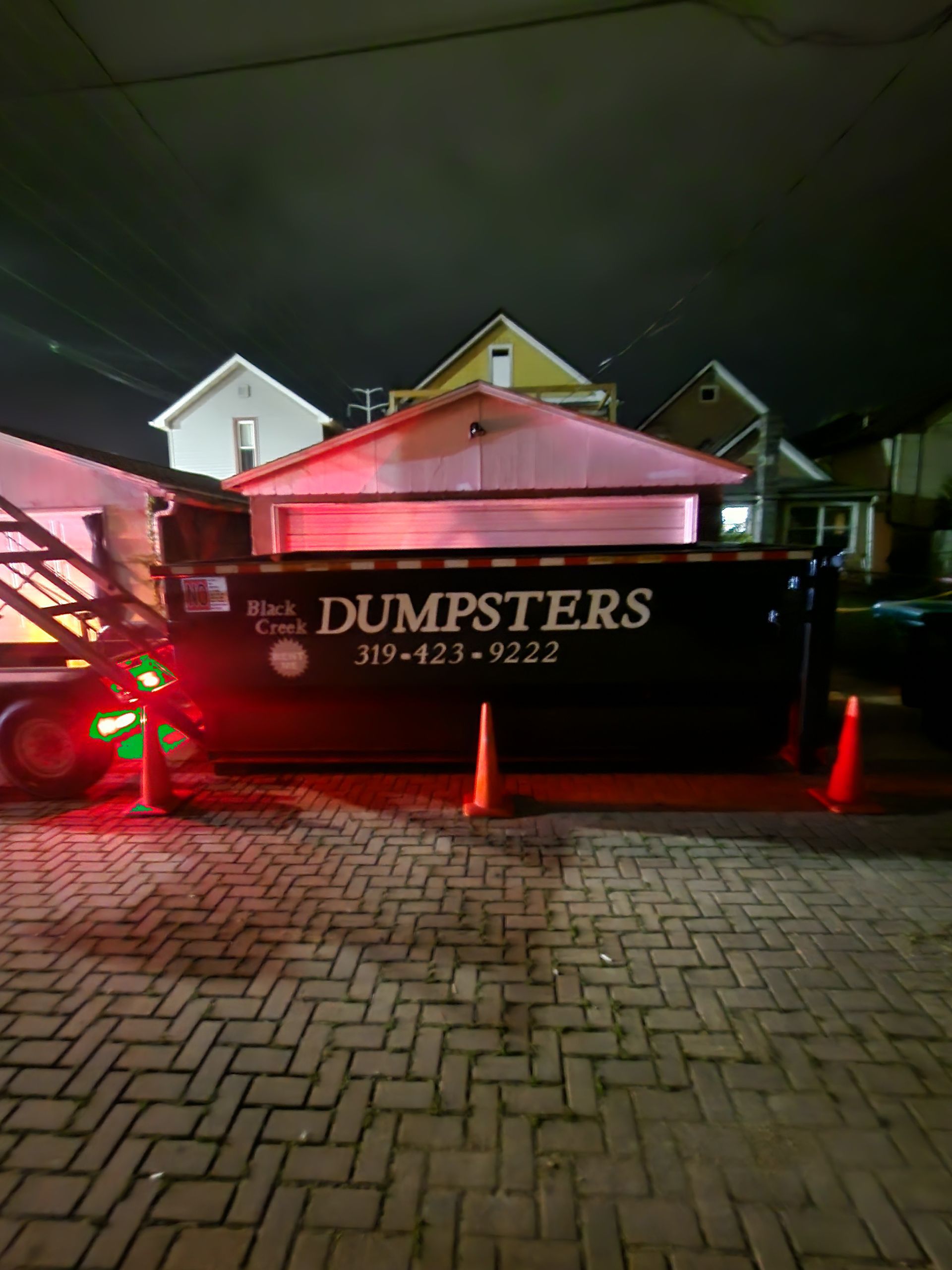 Black dumpster on a brick road at night with houses in the background.  Orange cones and a trailer nearby.