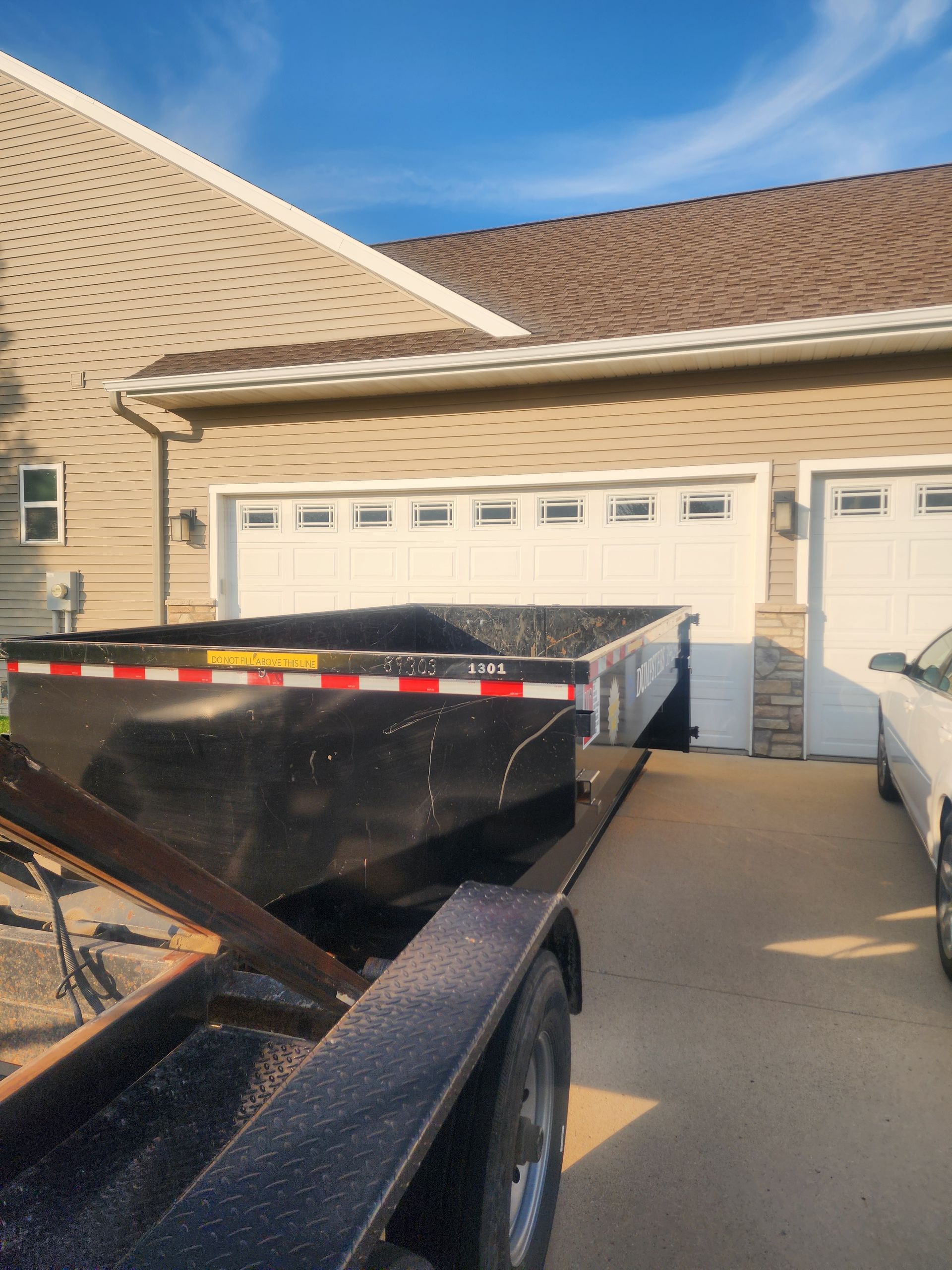 Dumpster on a trailer in front of a beige house with a brown roof and white garage doors.