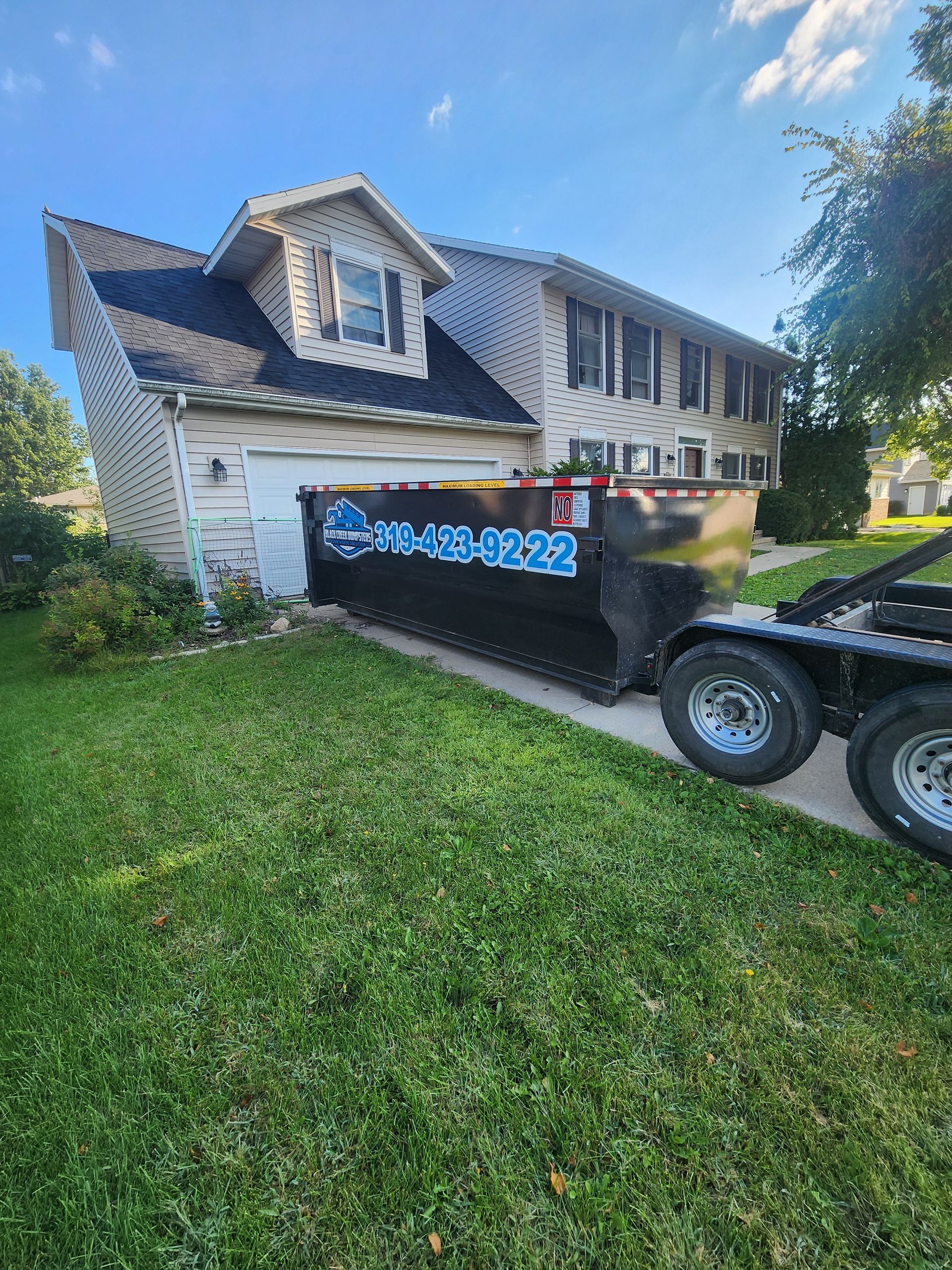 Black dumpster on a trailer parked in front of a two-story house with a green lawn.