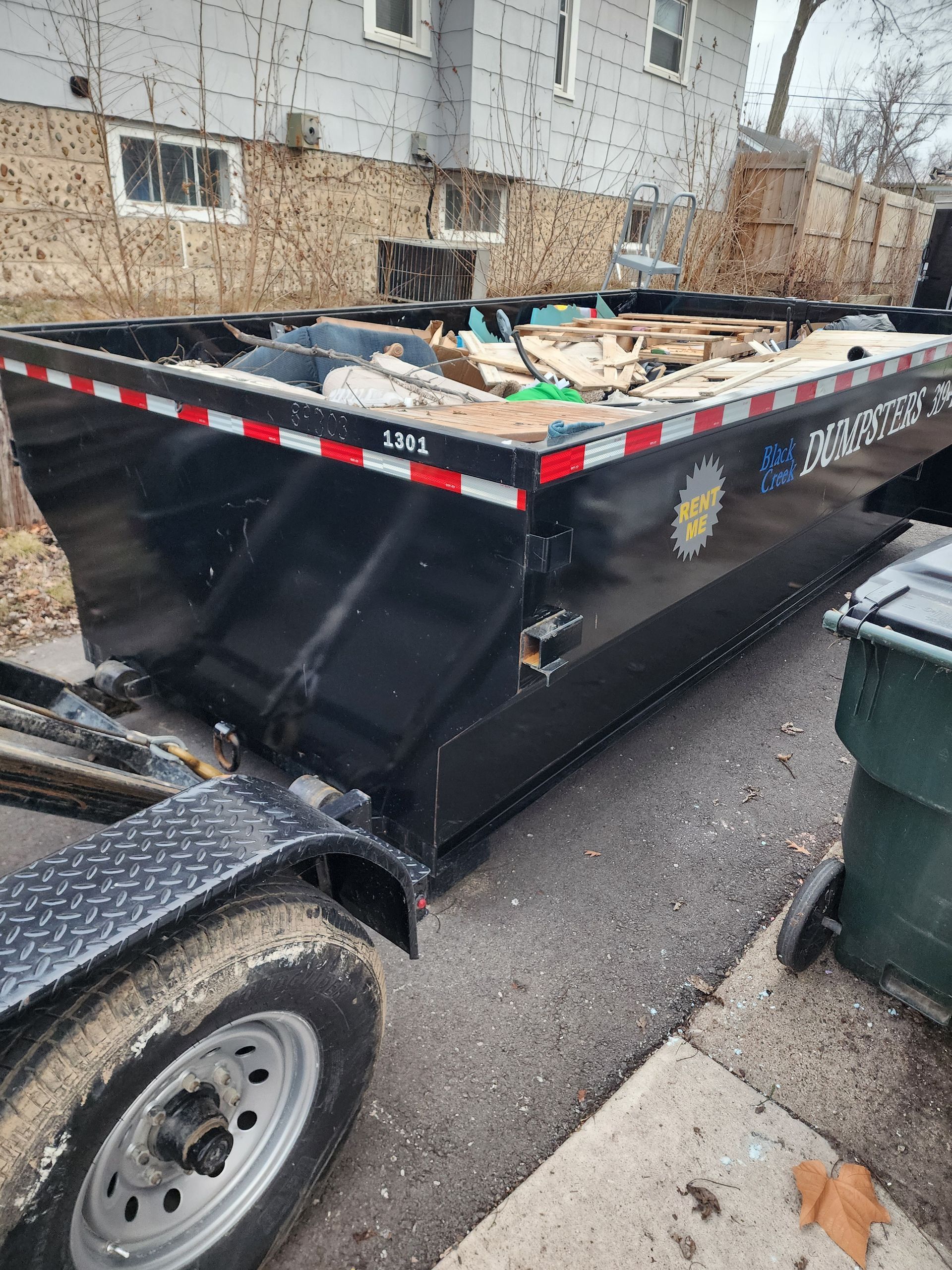Black dumpster on a trailer, filled with debris, parked on a paved surface near a building and a trash can.
