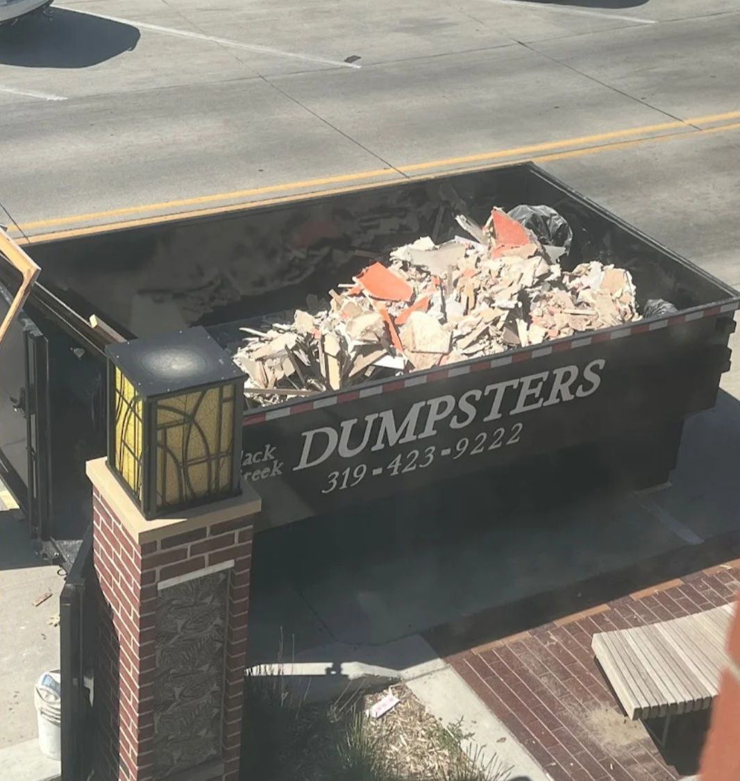A black dumpster filled with construction debris next to a brick column, with a street in the background.