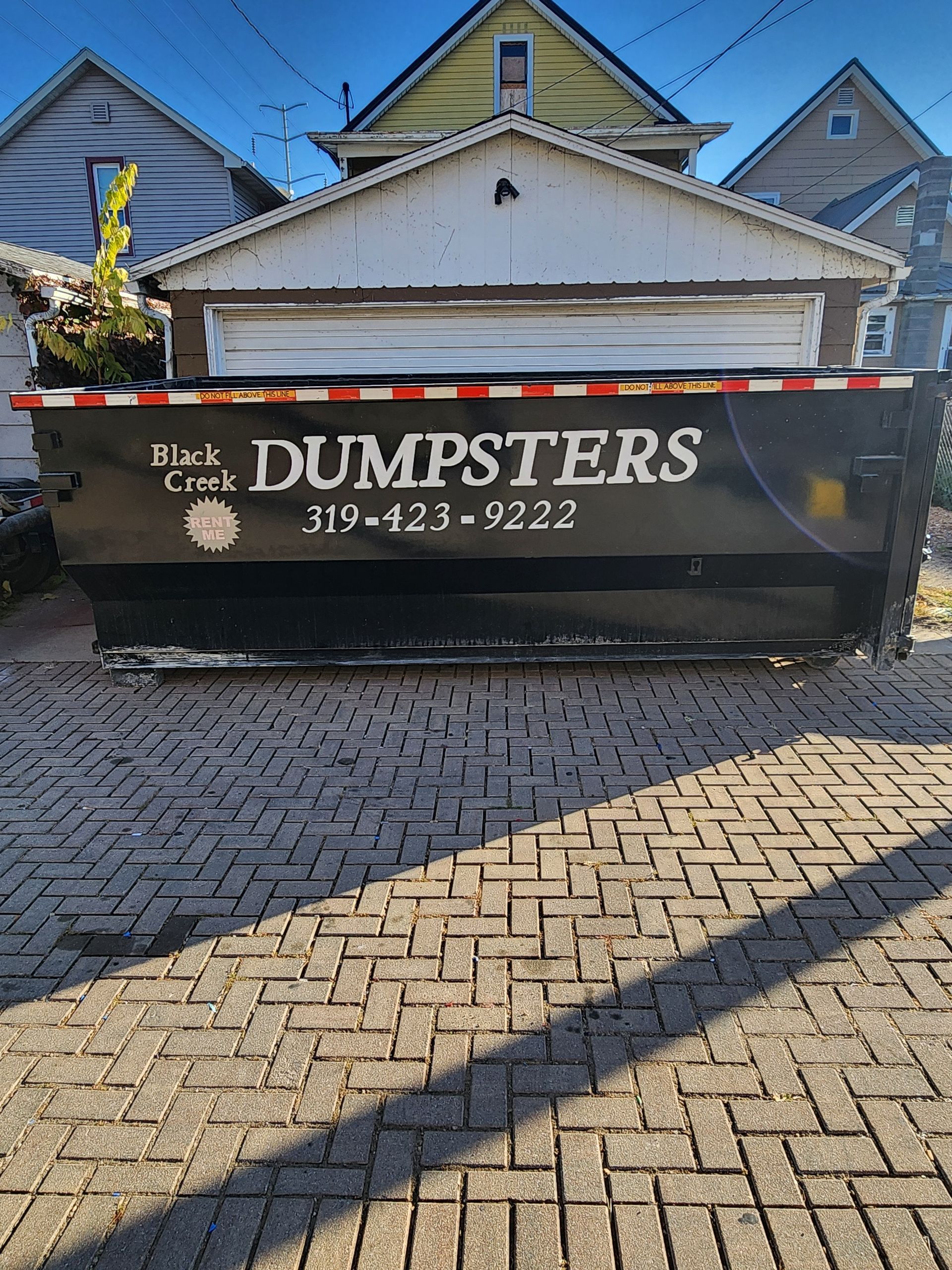 Black dumpster on a brick driveway, in front of a garage and houses.