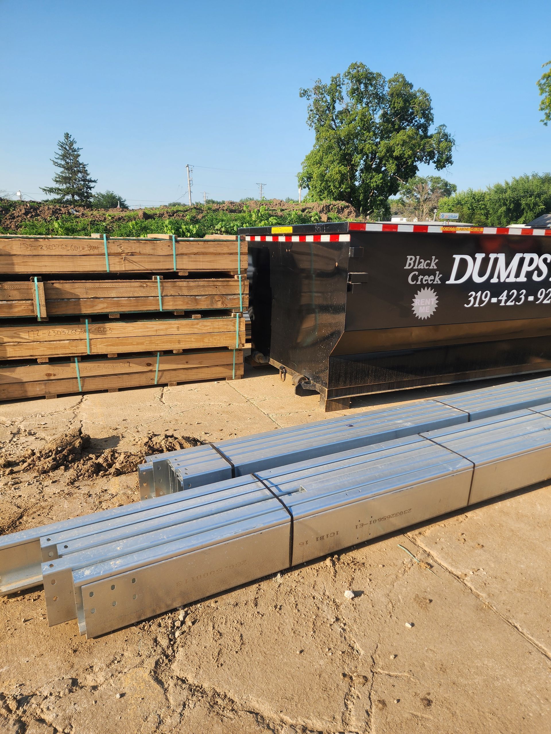 Construction site with dumpster, wood beams, and metal beams, on a sunny day.
