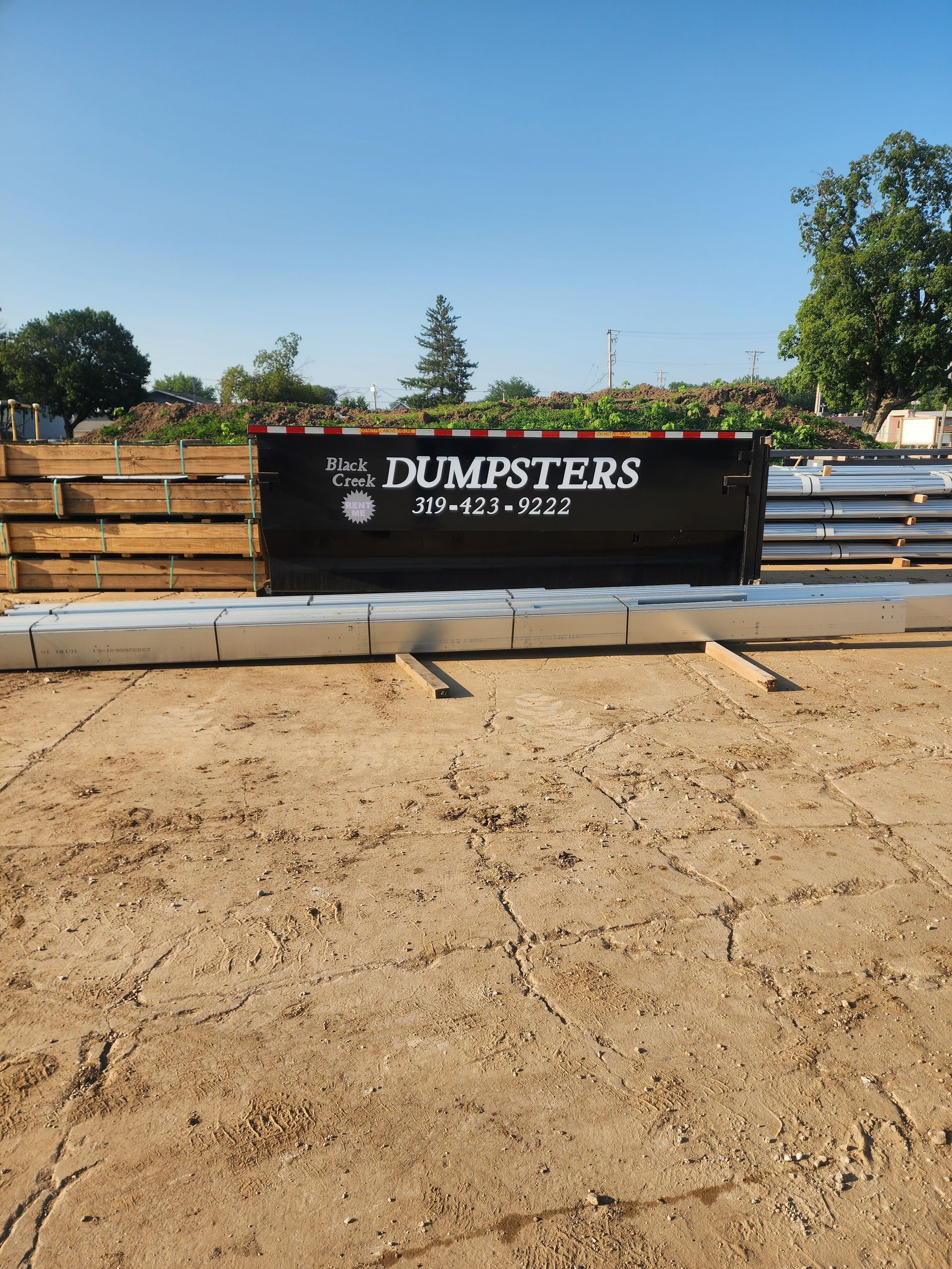 A black dumpster sits on cracked dirt, surrounded by wood and concrete.