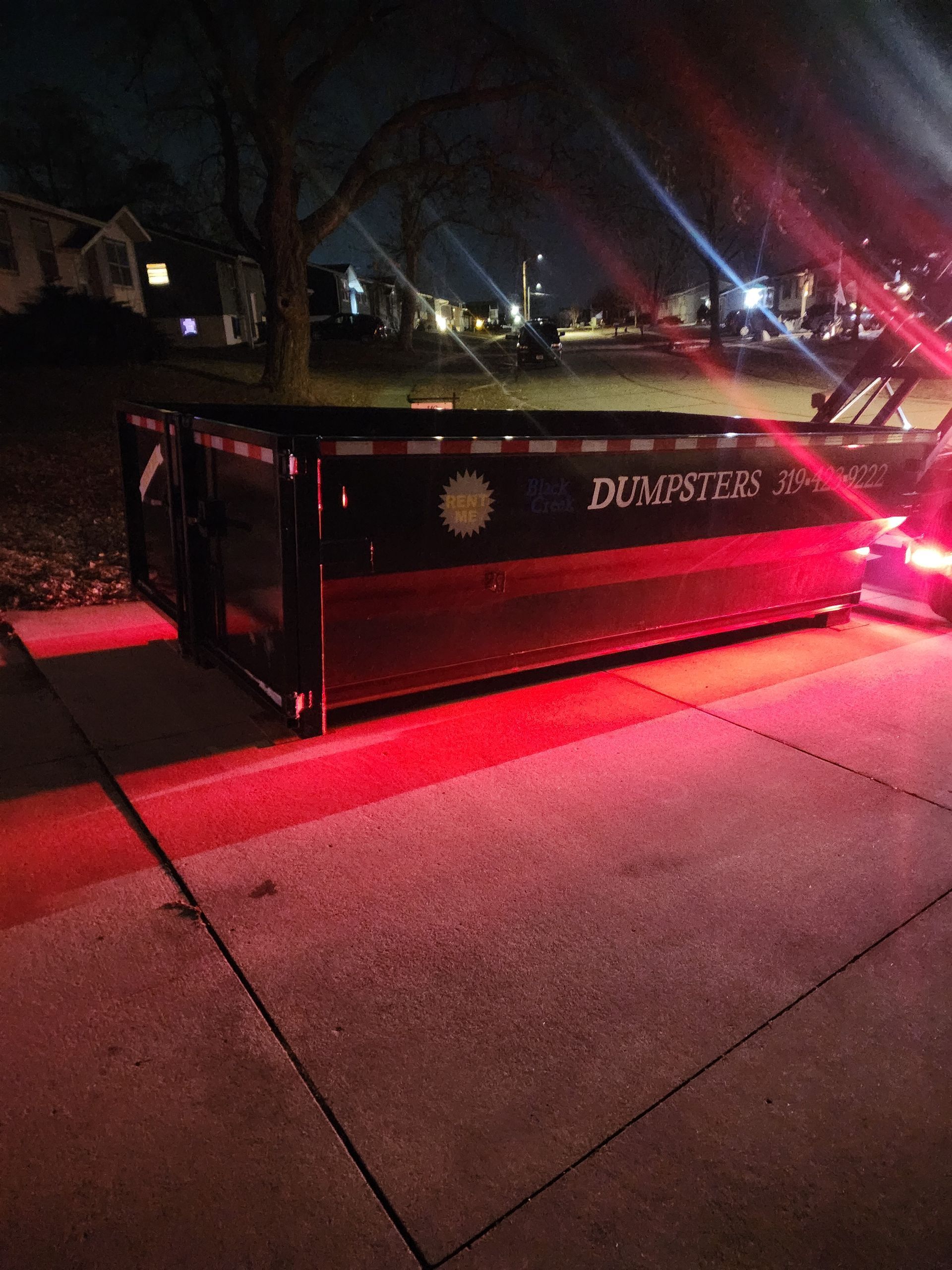 A black dumpster with red lights sits on a concrete driveway at night.