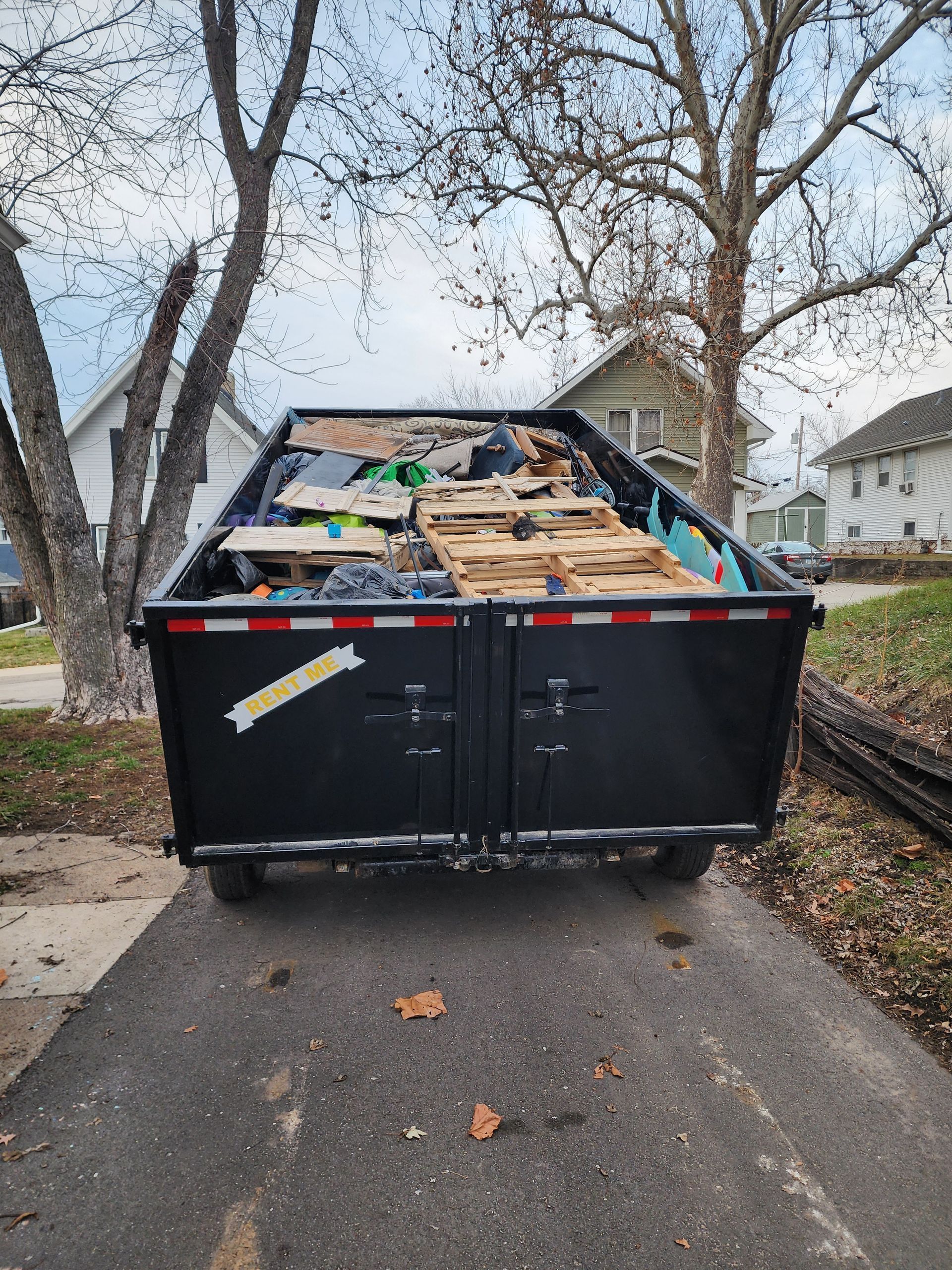 Black dumpster on a driveway filled with construction debris, trees and houses in background.