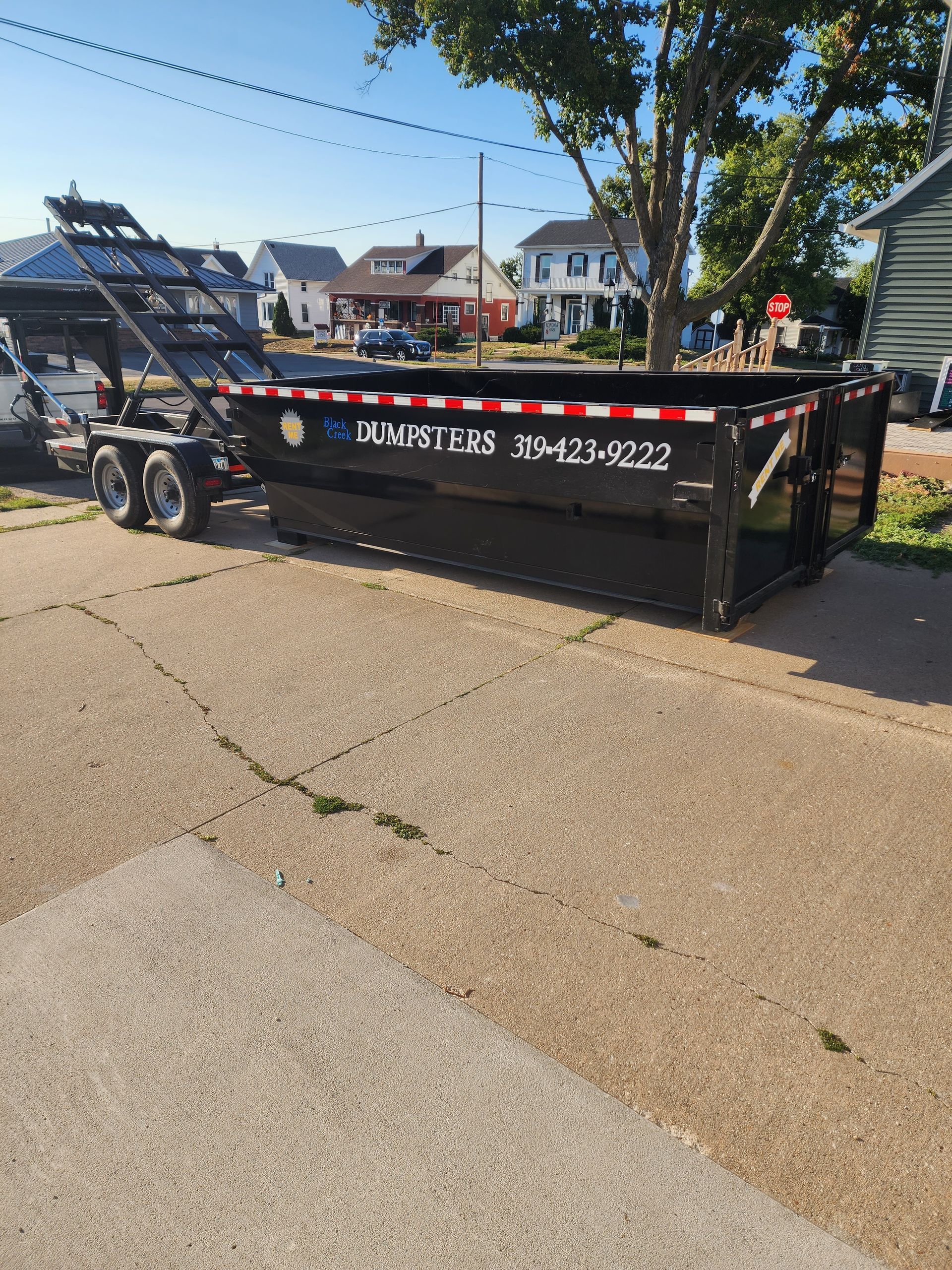Black dumpster on a trailer parked on a driveway, with houses in the background.