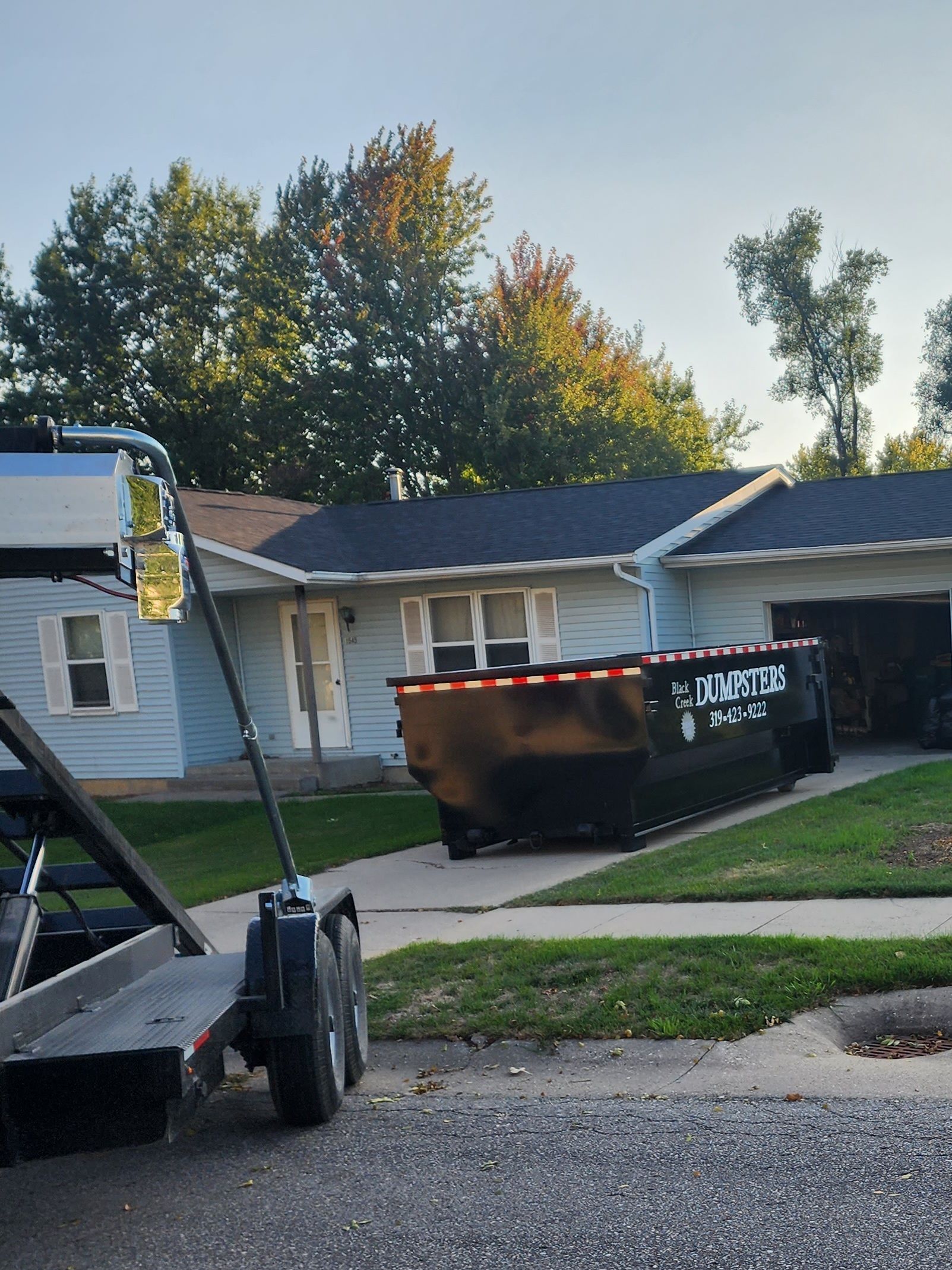 A black dumpster sits in a driveway in front of a blue house.