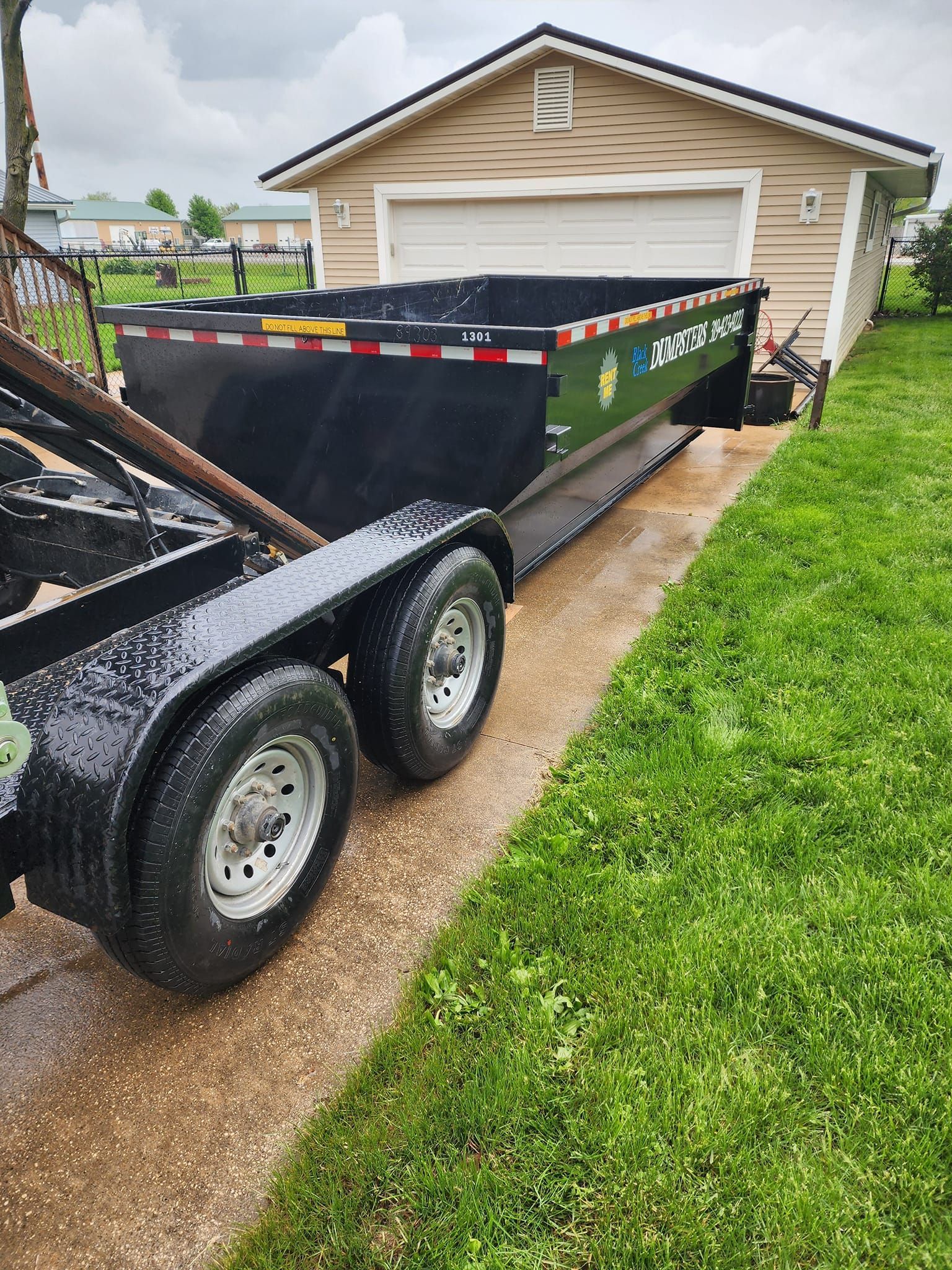 Black trailer with a dumpster parked next to a beige garage on a wet, grassy lawn.
