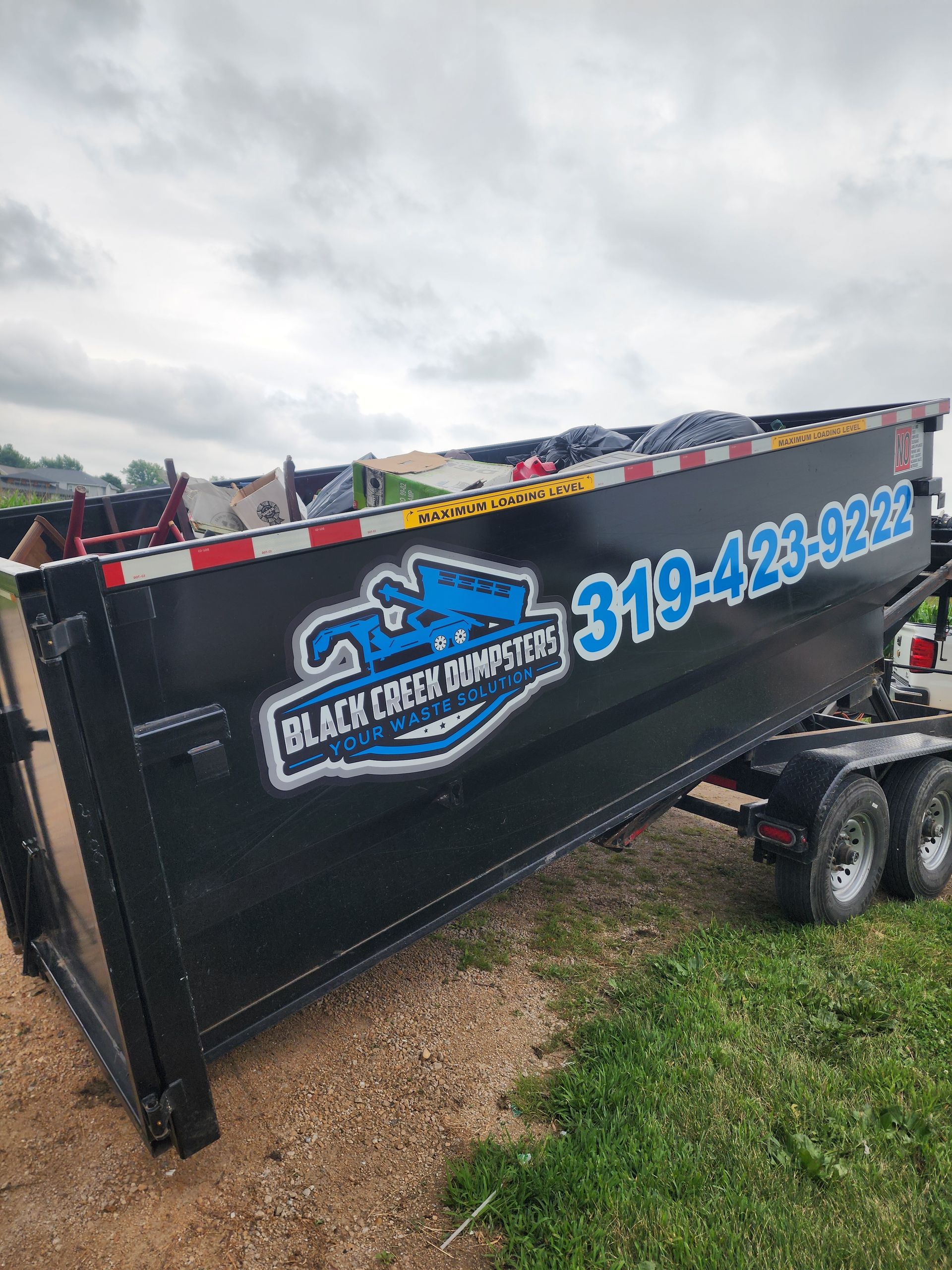 Black dumpster trailer with company logo, phone number, and a partially filled container against a cloudy sky.