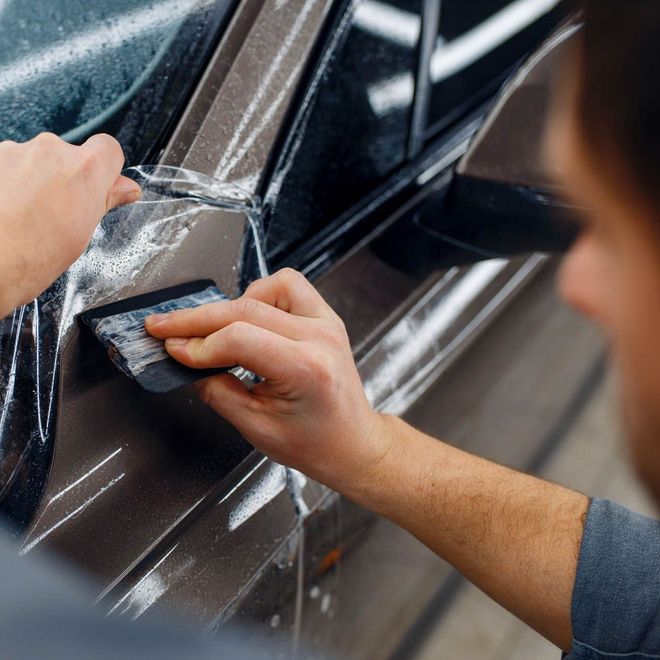 A person is using a blue squeegee to remove plastic from the hood of a car