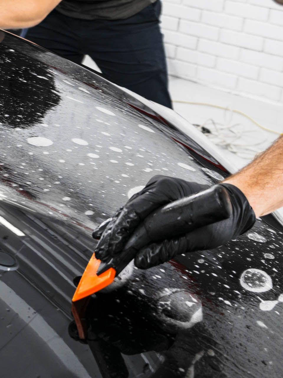 A person is using a blue squeegee to remove plastic from the hood of a car