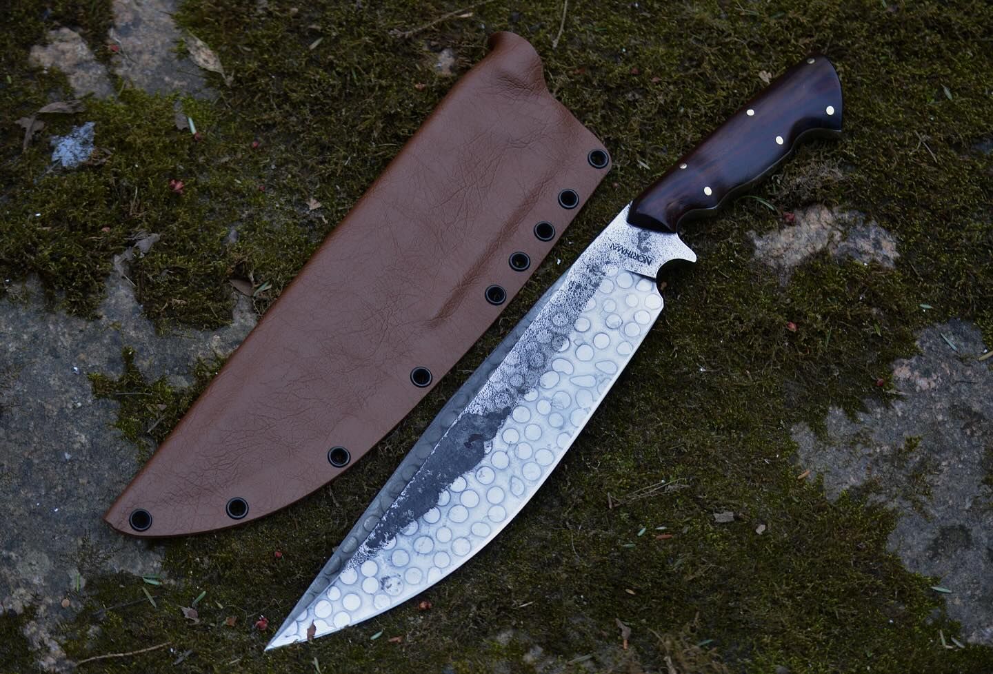 Large, rustic knife with a brown handle and textured blade, lying next to a brown leather sheath on moss.