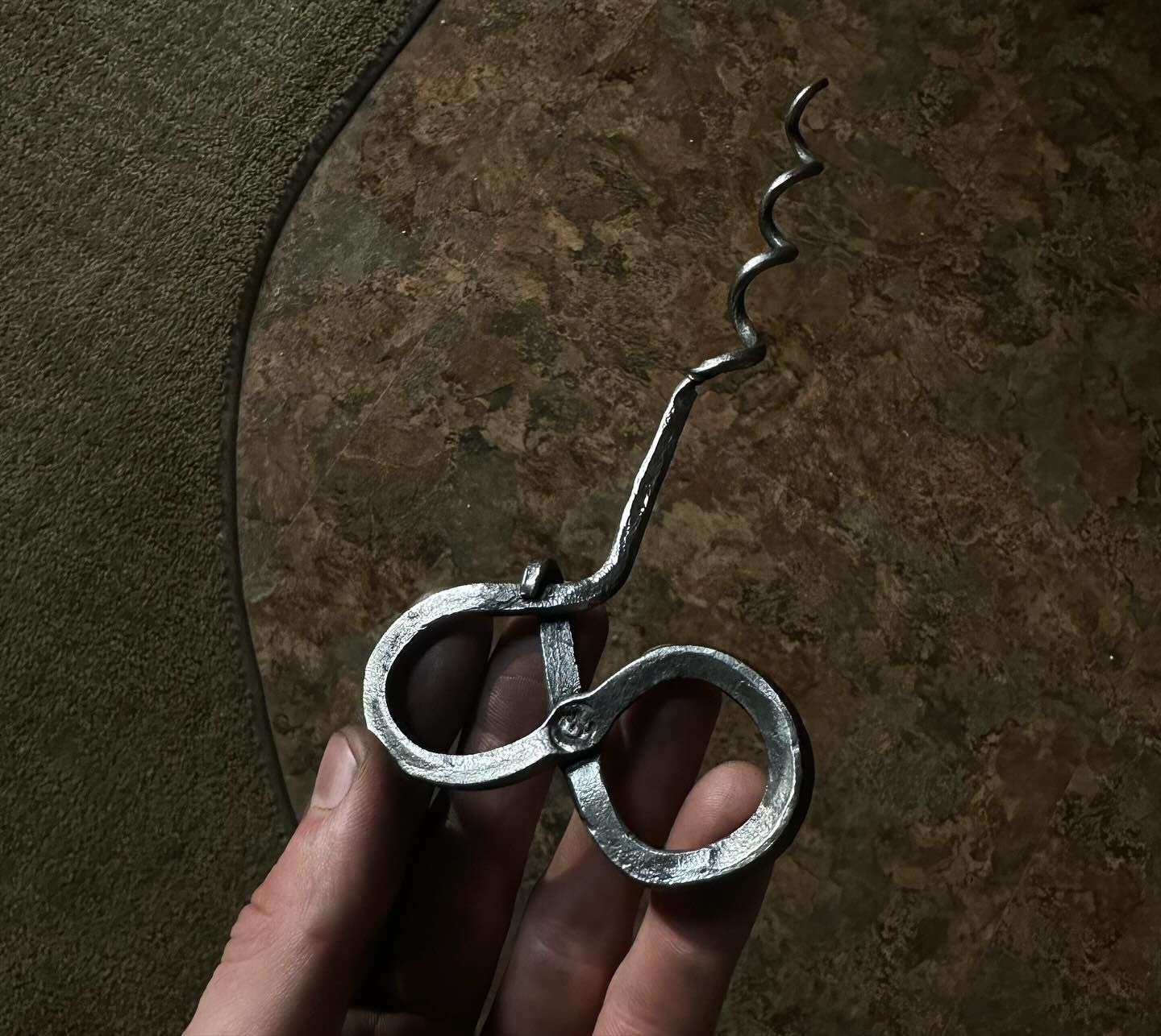Hand holding a hand-forged metal corkscrew with two loop handles and a spiraled worm.