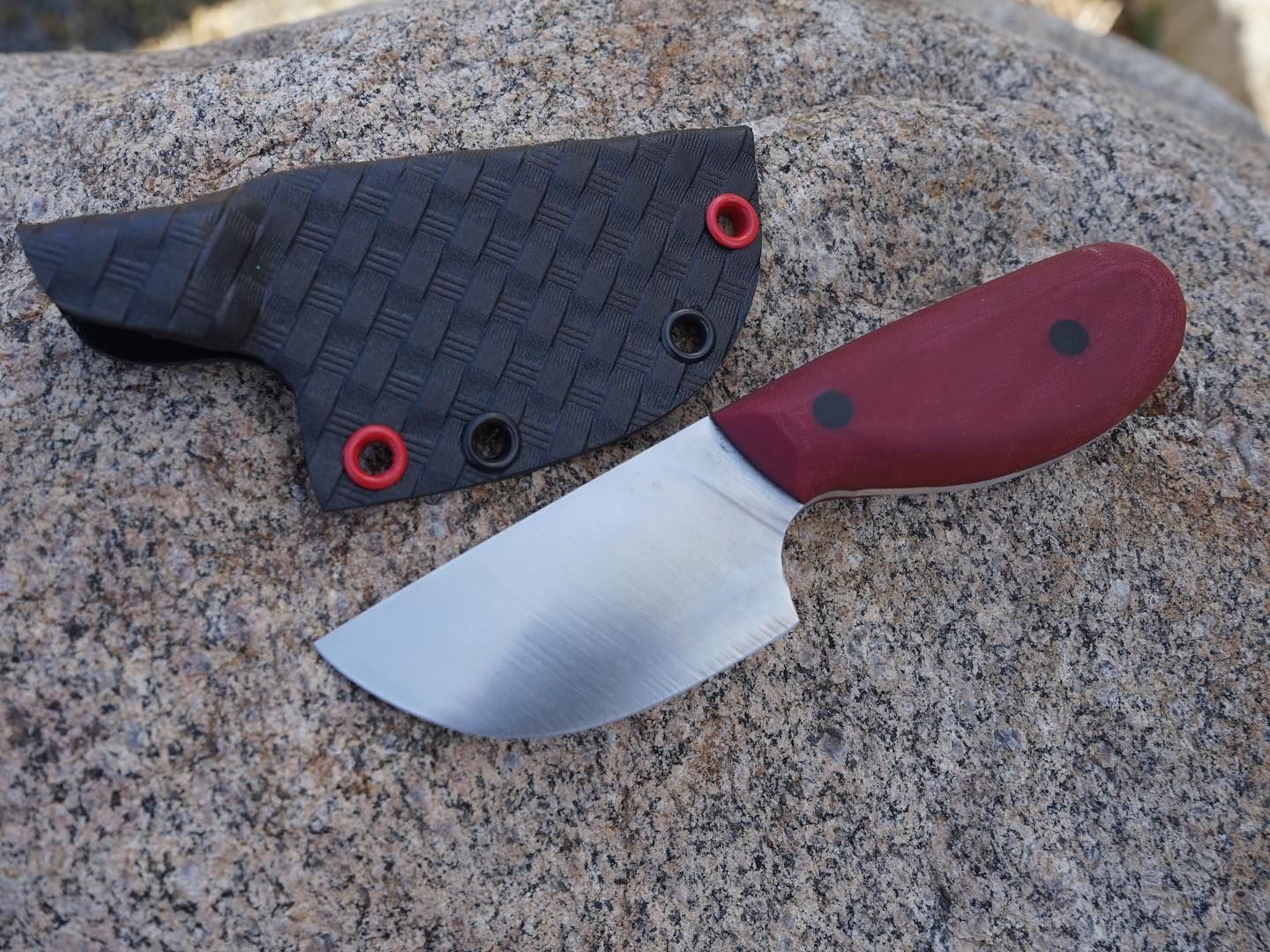 Small burgundy-handled knife with silver blade and black sheath on a rock.