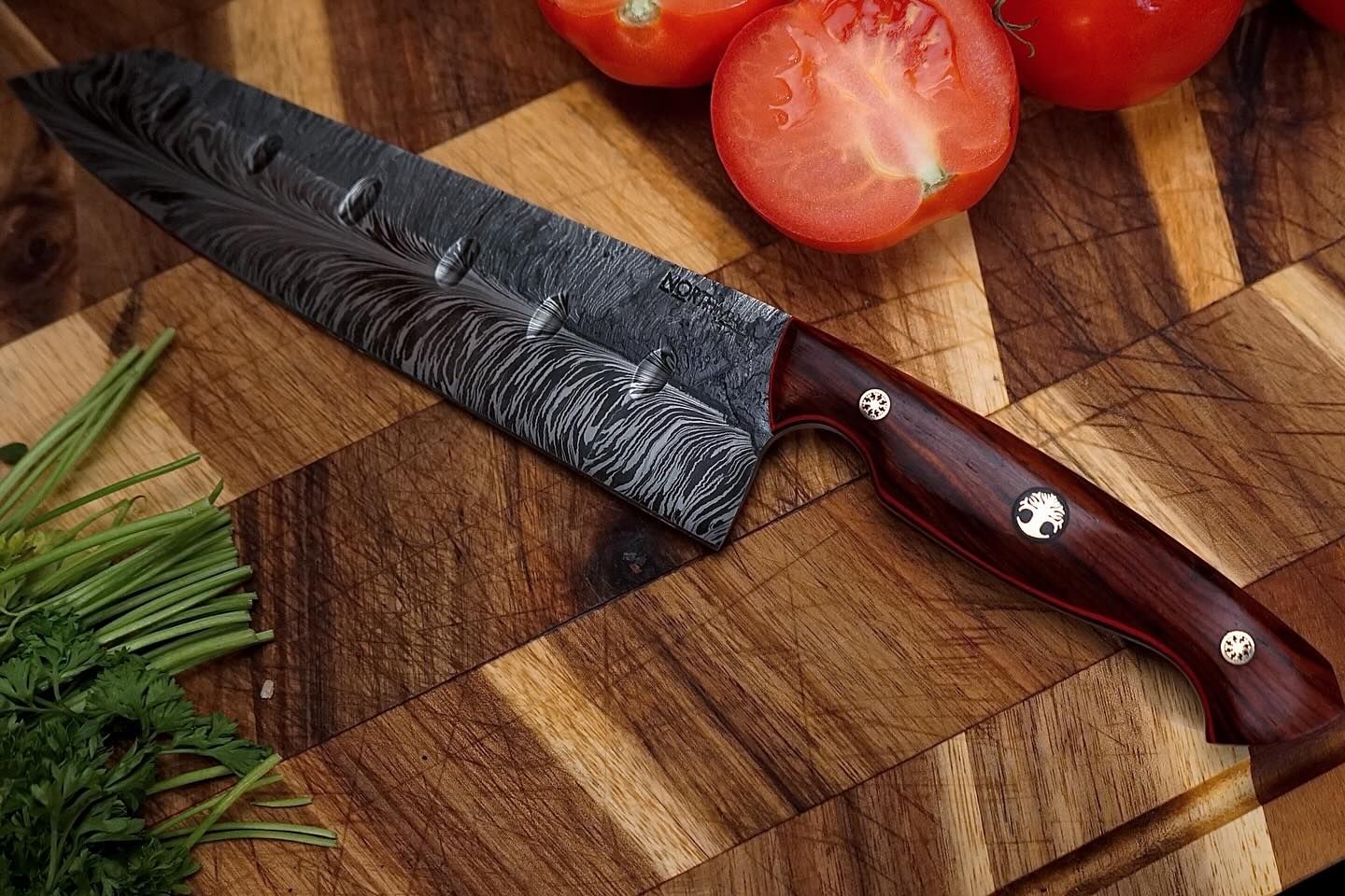 Damascus steel chef's knife on a wooden cutting board with tomatoes and parsley.