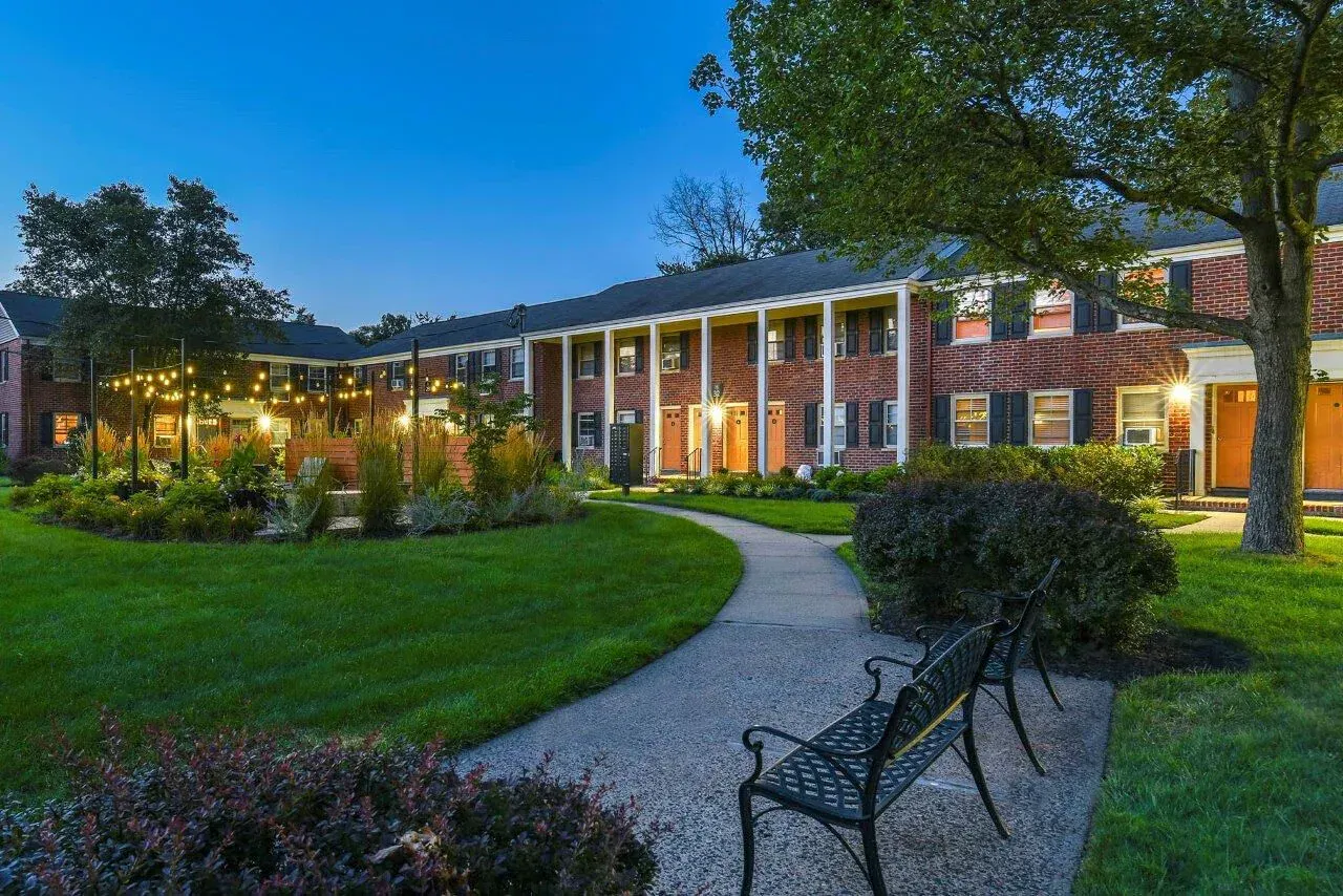 Exterior view of a brick apartment building with a curved path, benches, and string lights in a landscaped courtyard.