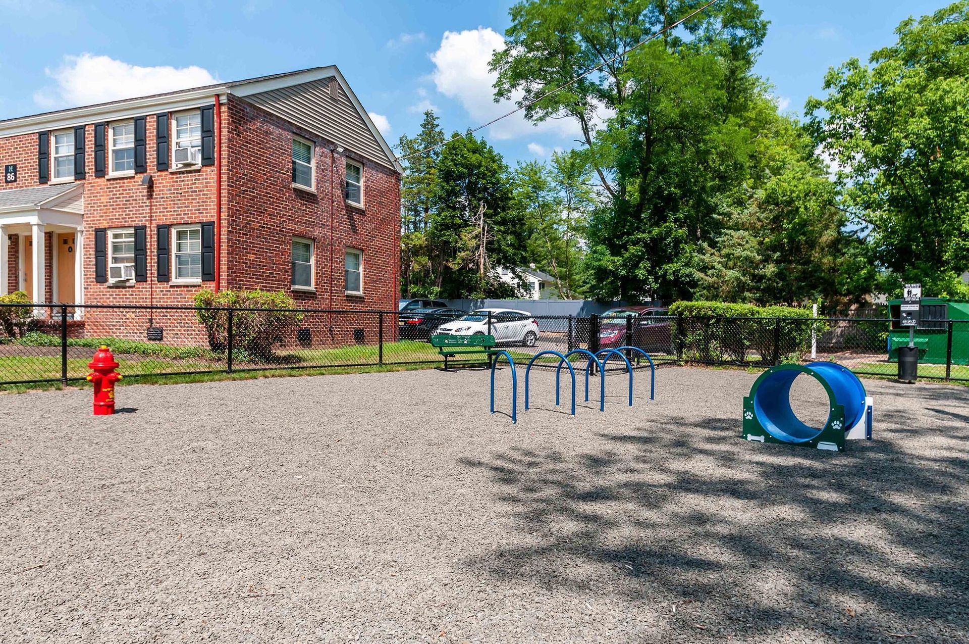 Gravel playground area with blue arch bars and a blue tunnel beside a brick apartment building, fenced.