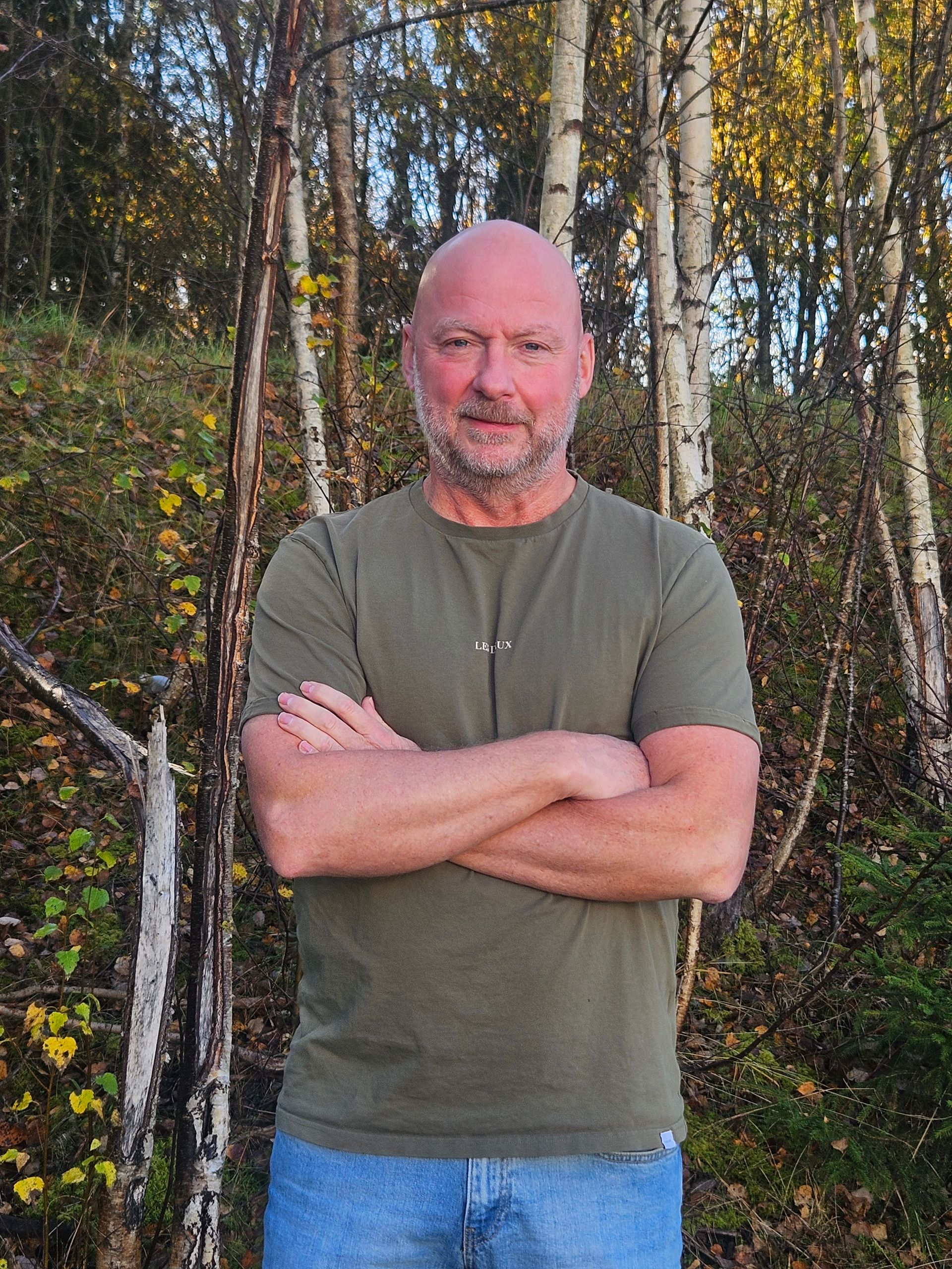 Mann med armene i kors, iført grønn skjorte og jeans, står utendørs i nærheten av trær med hvit bark.