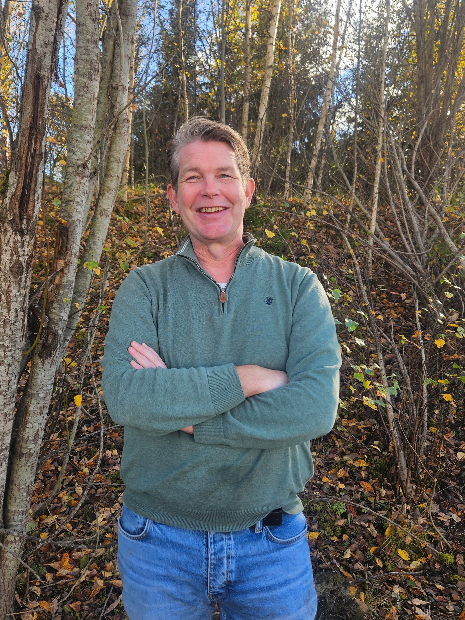 Mann med armene i kors, smilende, iført en grønn genser og jeans, står utendørs i nærheten av trær med gule blader.