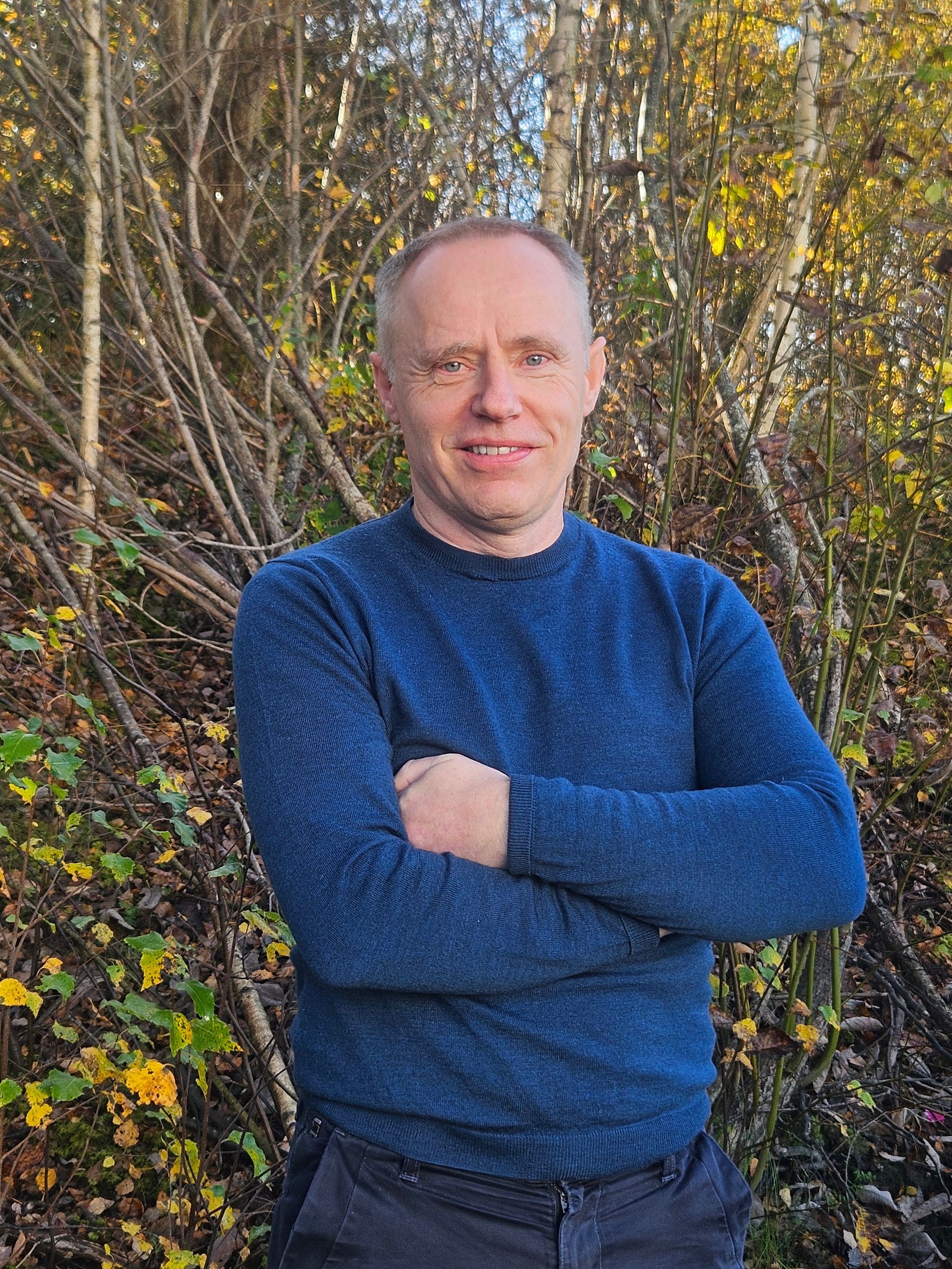 Mann i blå genser med armene i kors står foran trær.