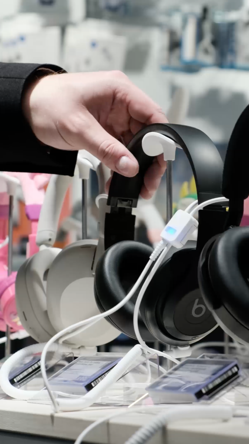 En hånd som holder svarte hodetelefoner i en butikkutstilling med synlige hvite og rosa hodetelefoner og prislapper.