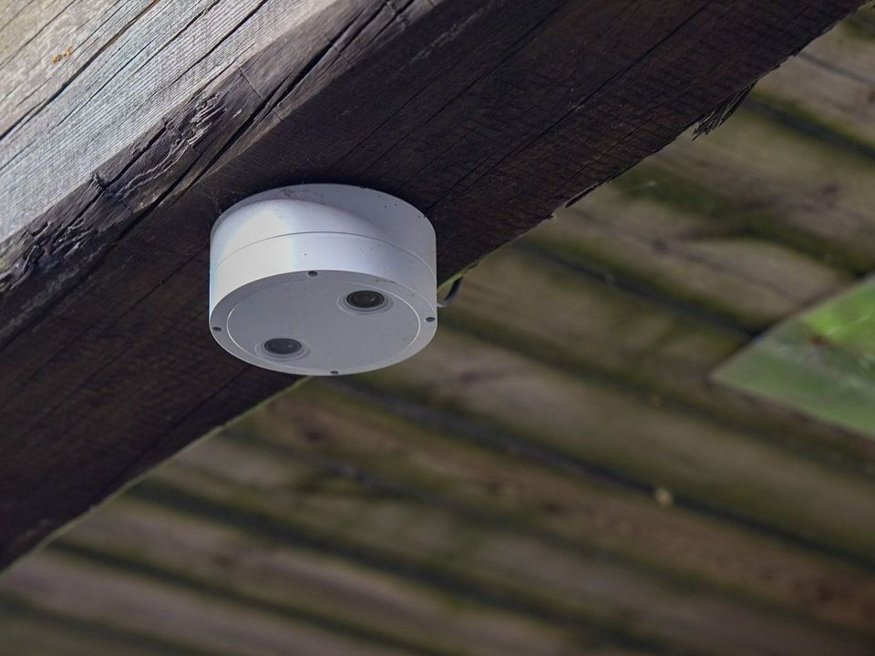 Hvitt, sylindrisk sikkerhetskamera montert på en mørk trebjelke, vendt nedover.