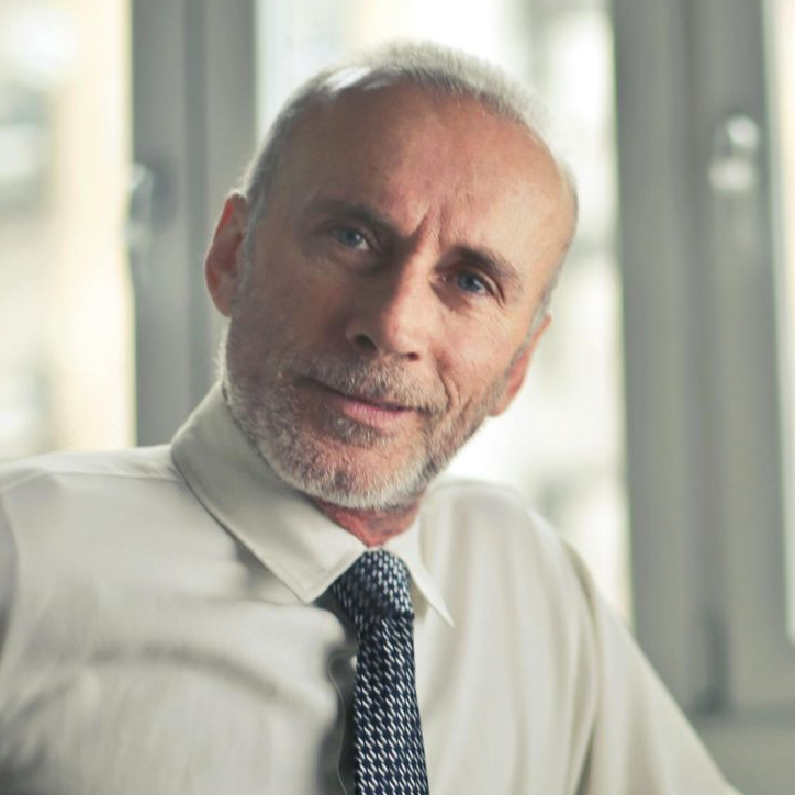 Mann i hvit skjorte og mønstret slips, ser på betrakteren med et lite smil, innendørs.