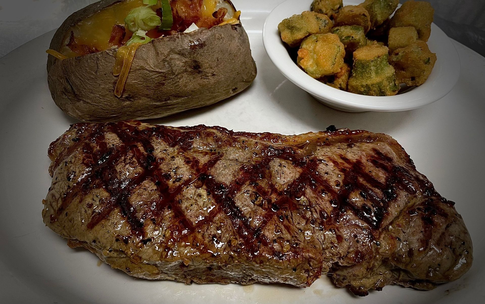 A steak with a baked potato and a bowl of broccoli on a white plate.