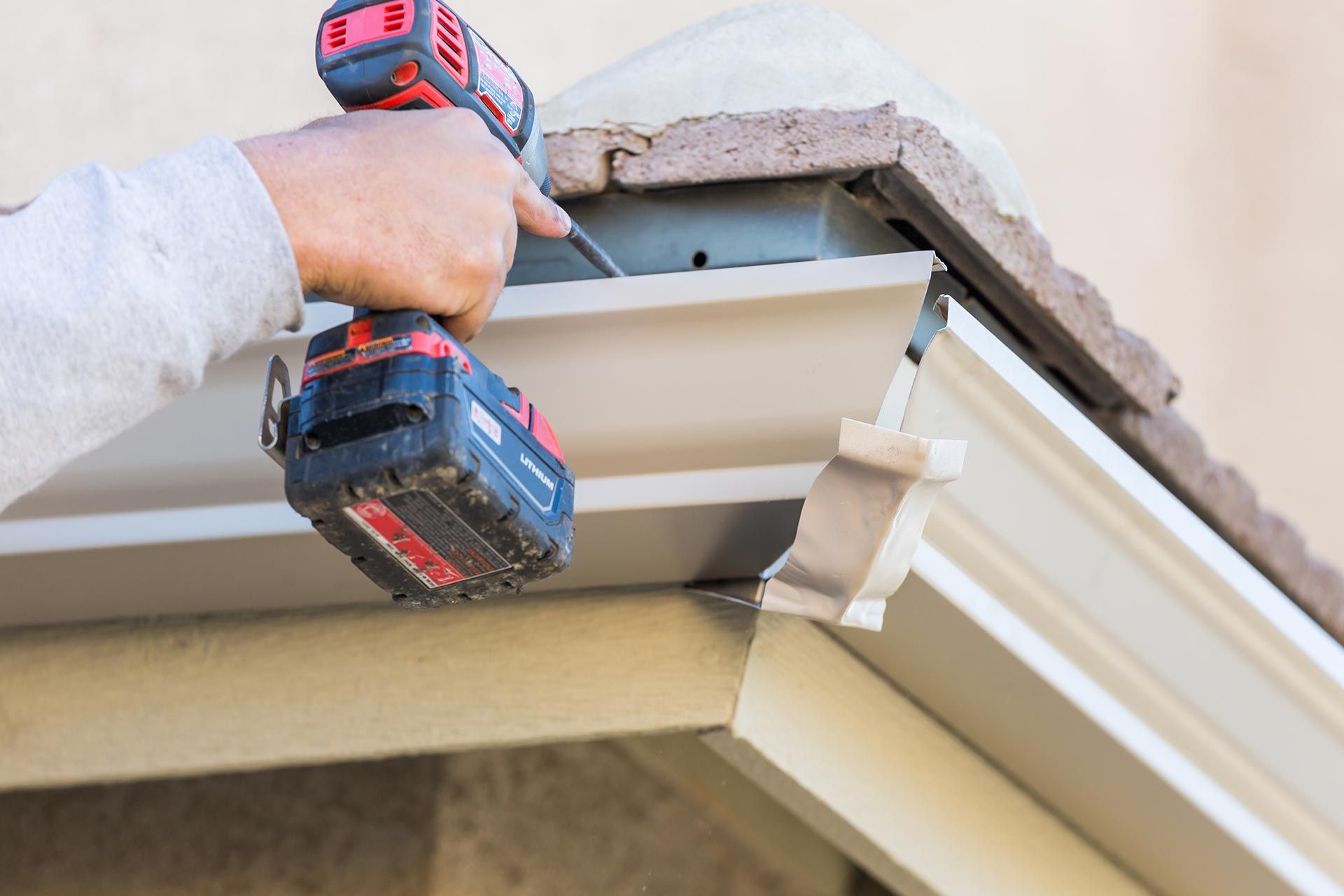 Worker installing a white gutter with a red-and-black drill on a roof edge Worker installing a white gutter with a red-and-black drill on a roof edge
