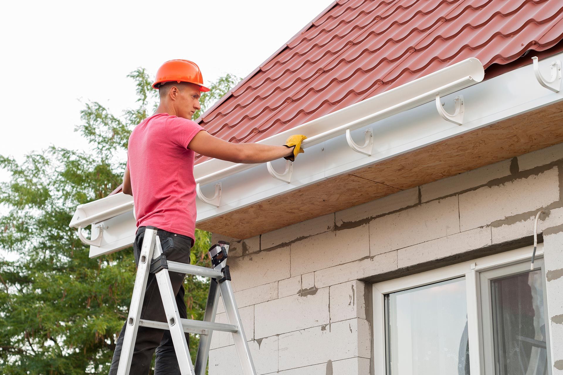 Worker on a ladder painting a house gutter under a red-tiled roof