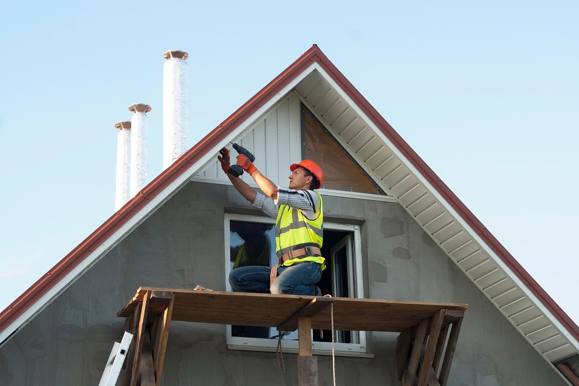 Worker on a roof using a power tool beside a house gable, wearing a safety vest and hard hat.