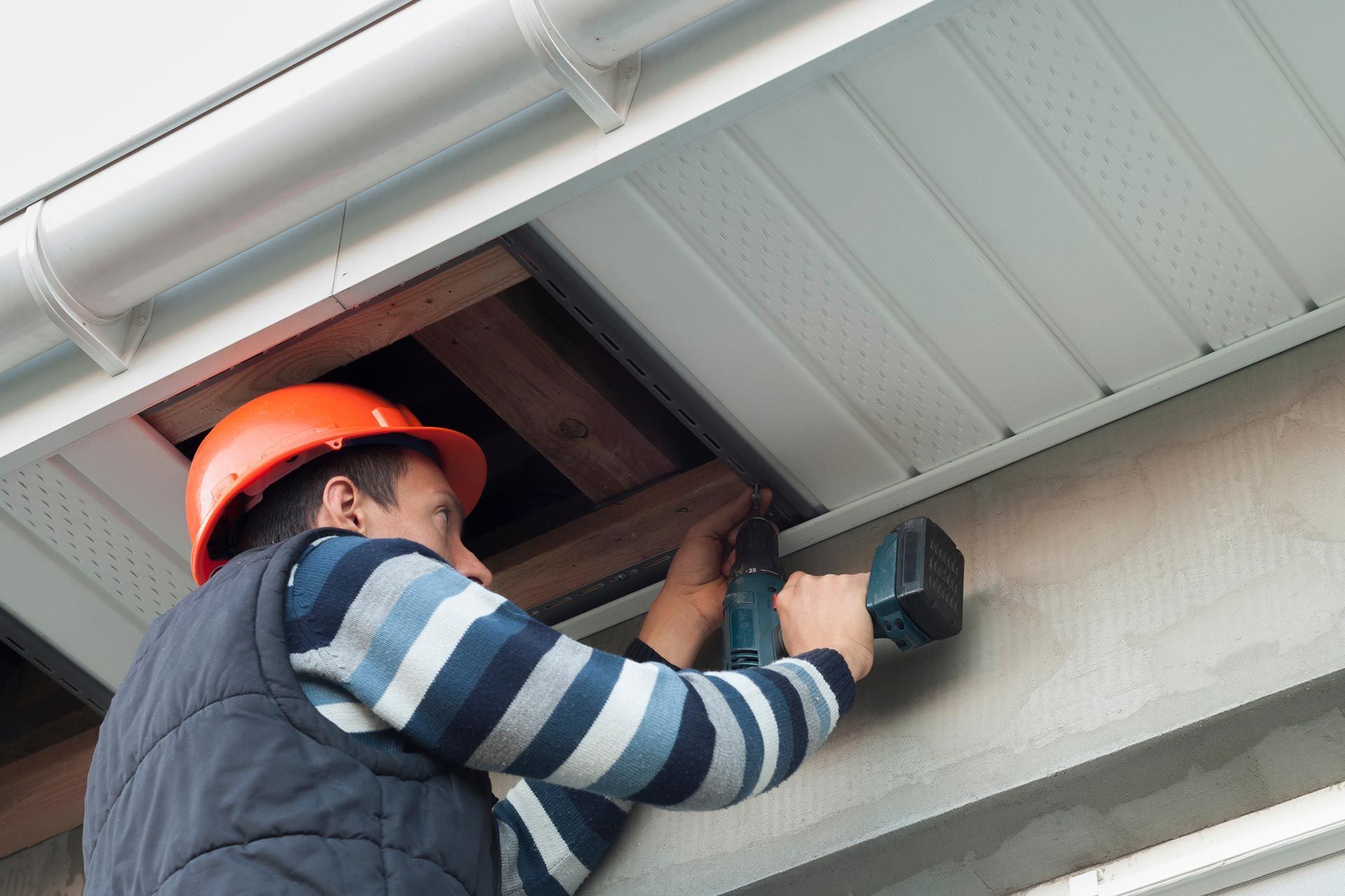 Worker in orange hard hat using a power drill beneath a roof overhang