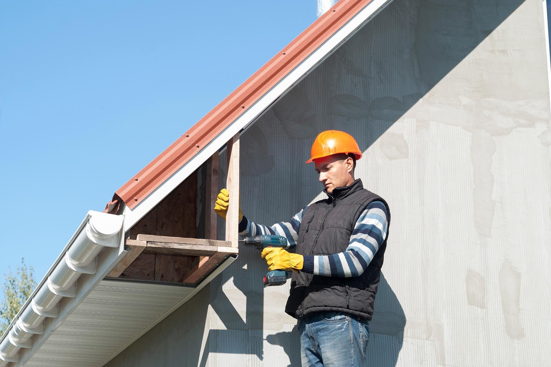 Worker in orange hard hat inspecting gutter on a house roof under clear blue sky