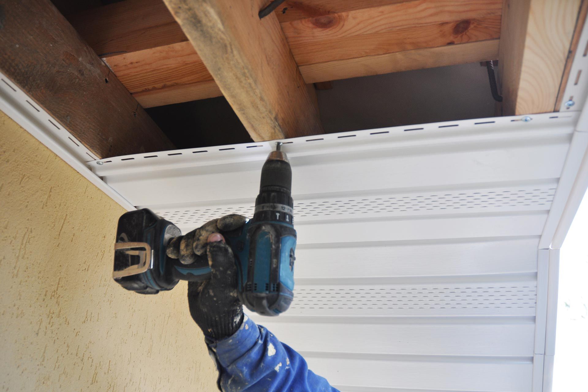 Worker using a blue drill to install screws in a white soffit under exposed wooden beams