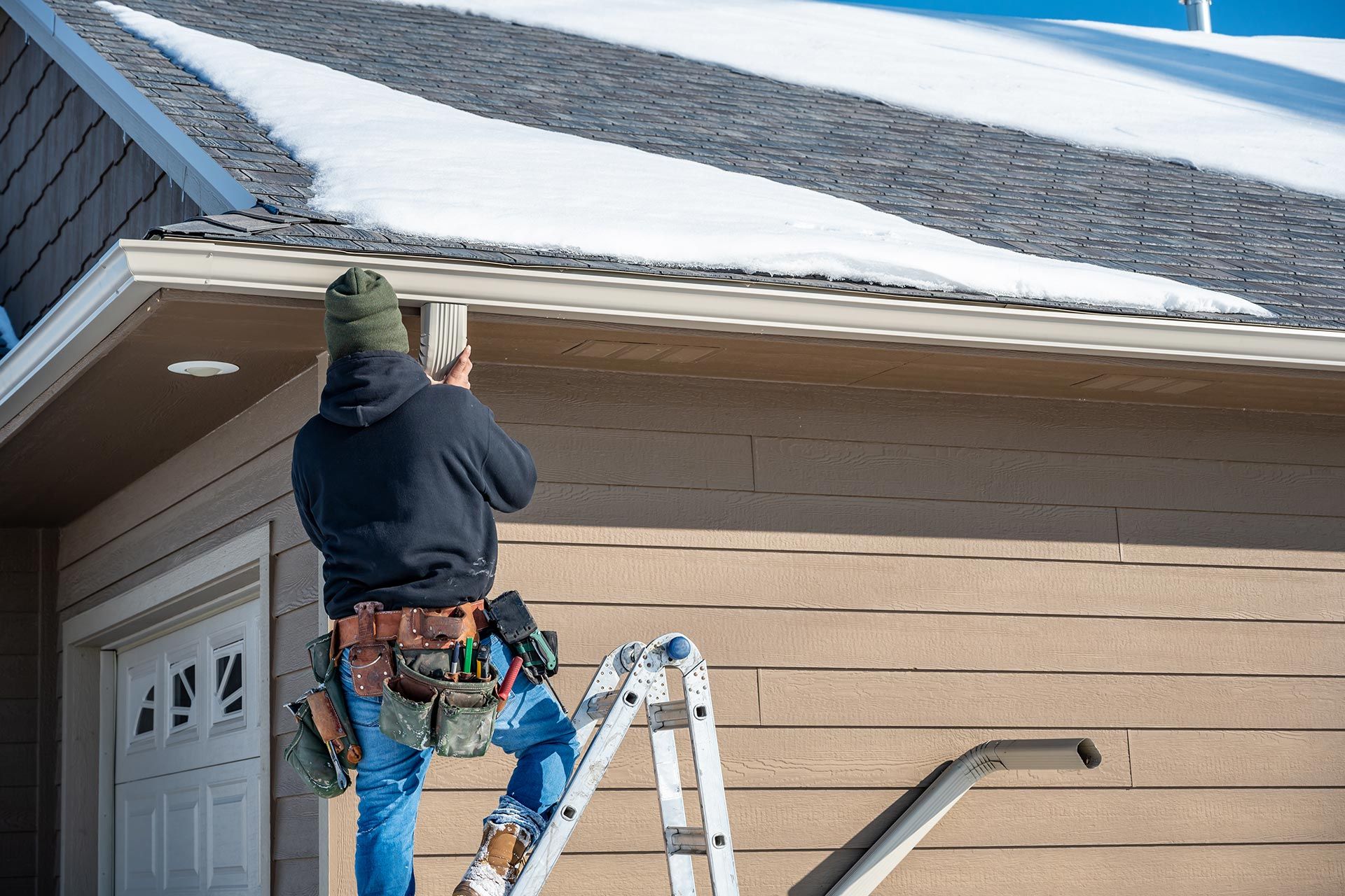 Worker on a ladder repairing white trim on a tan house roofline in winter Worker on a ladder repairing white trim on a tan house roofline in winter