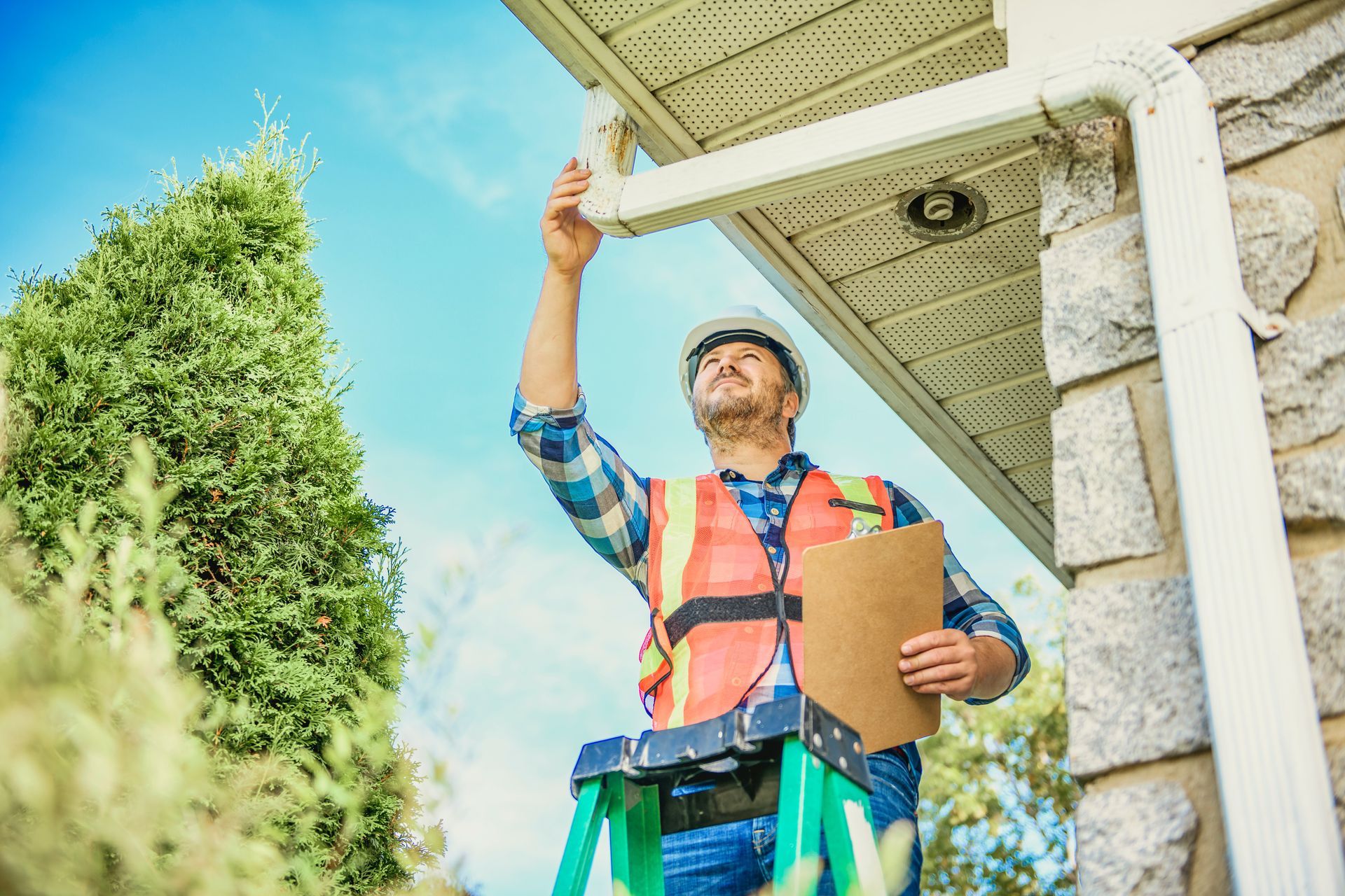 Worker in a hard hat inspecting a roof gutter with clipboard at a house exterior