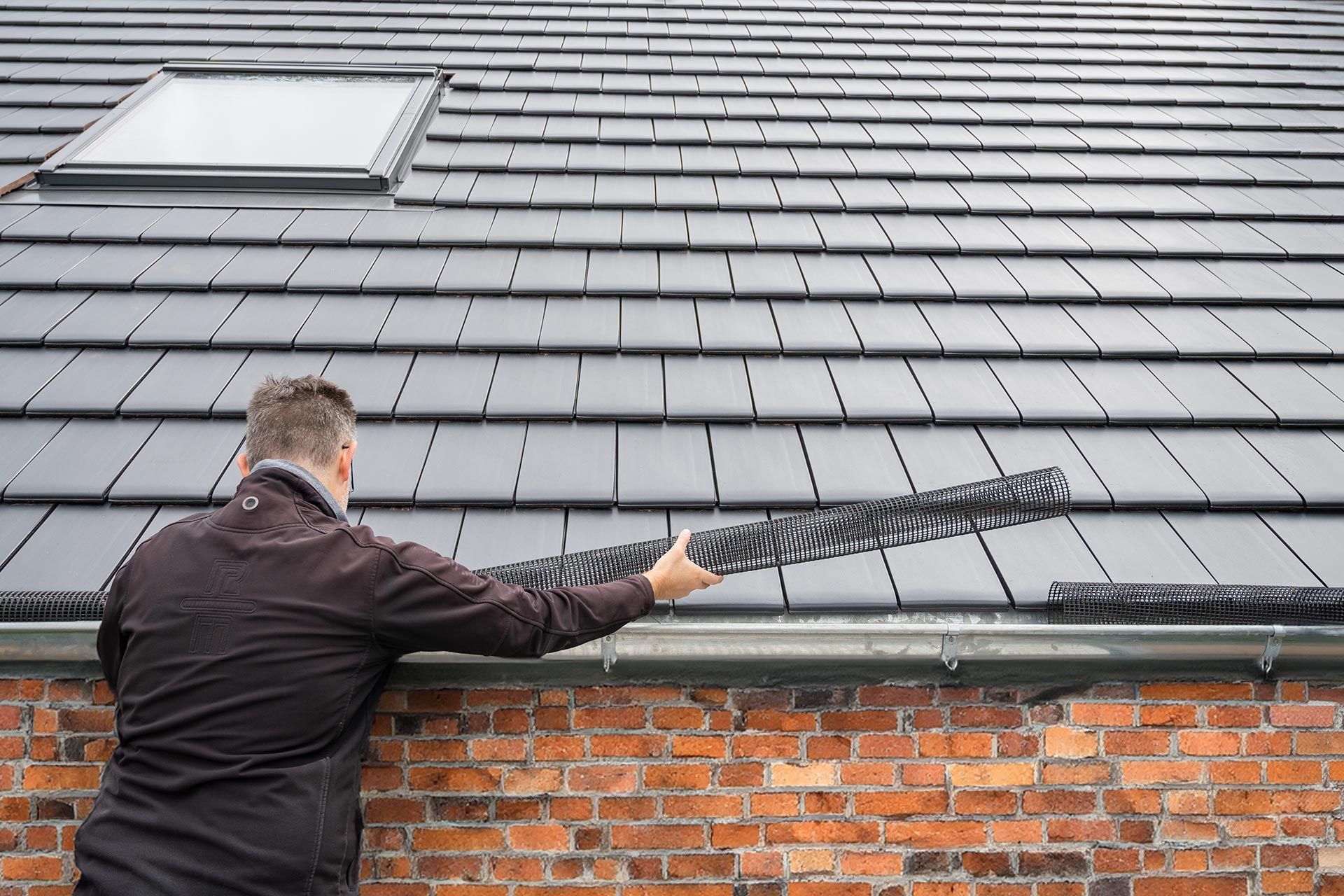 Person using a pressure washer to clean a gray roof above a brick wall