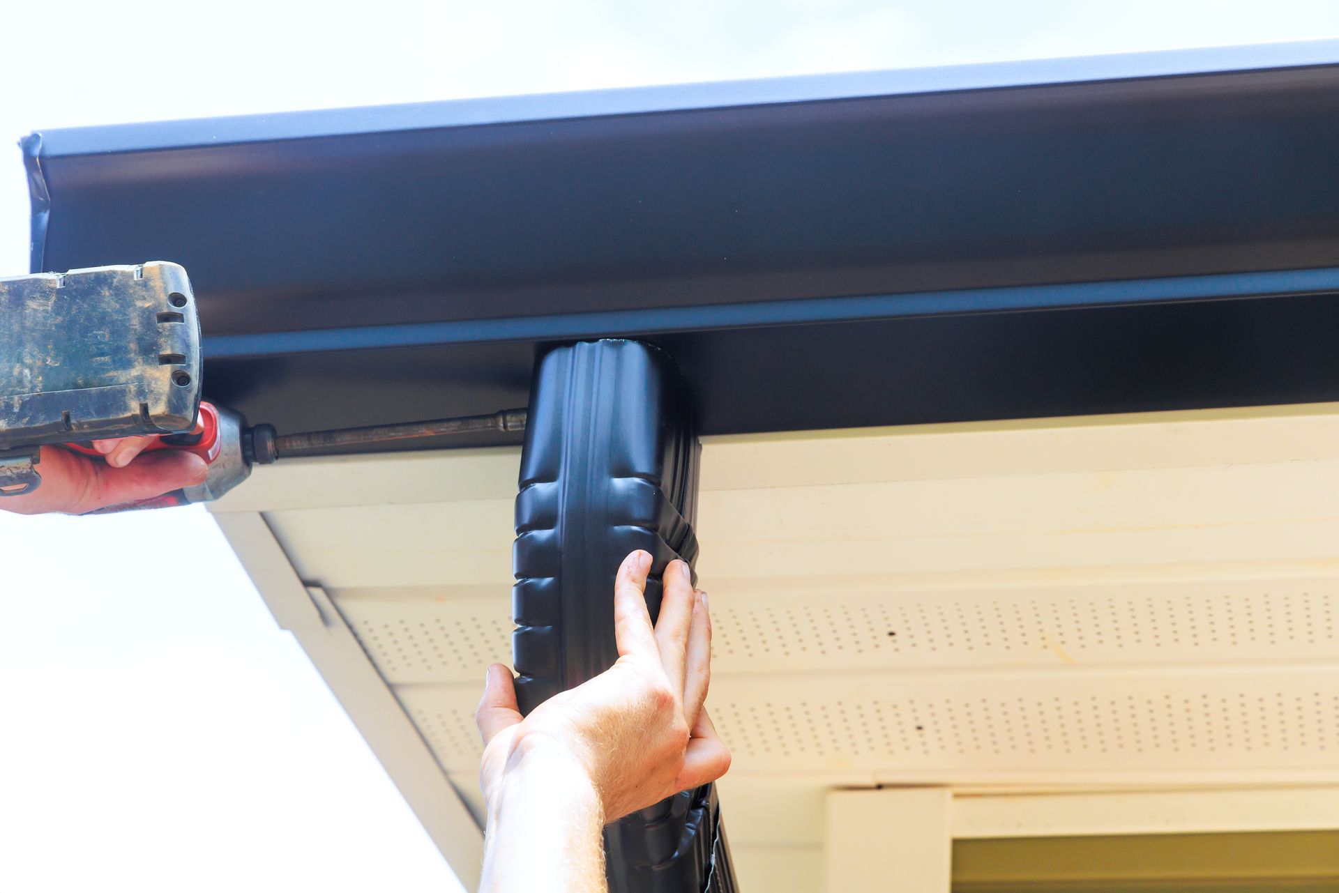 Hand holding a black strap under a roof eave next to a gutter or awning edge