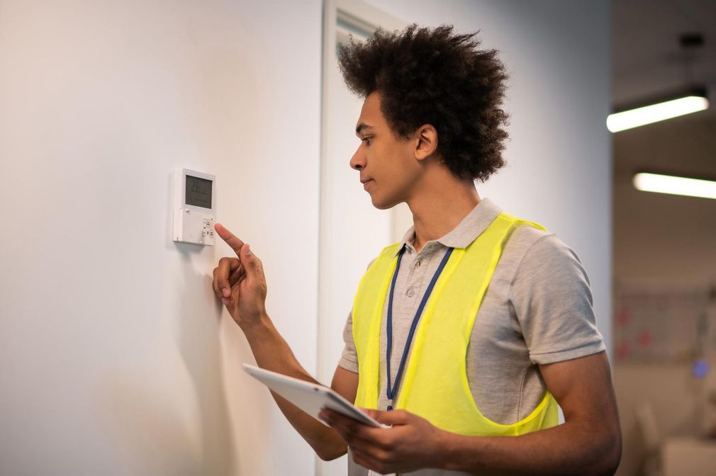 A man in a yellow vest is adjusting a thermostat while holding a tablet.