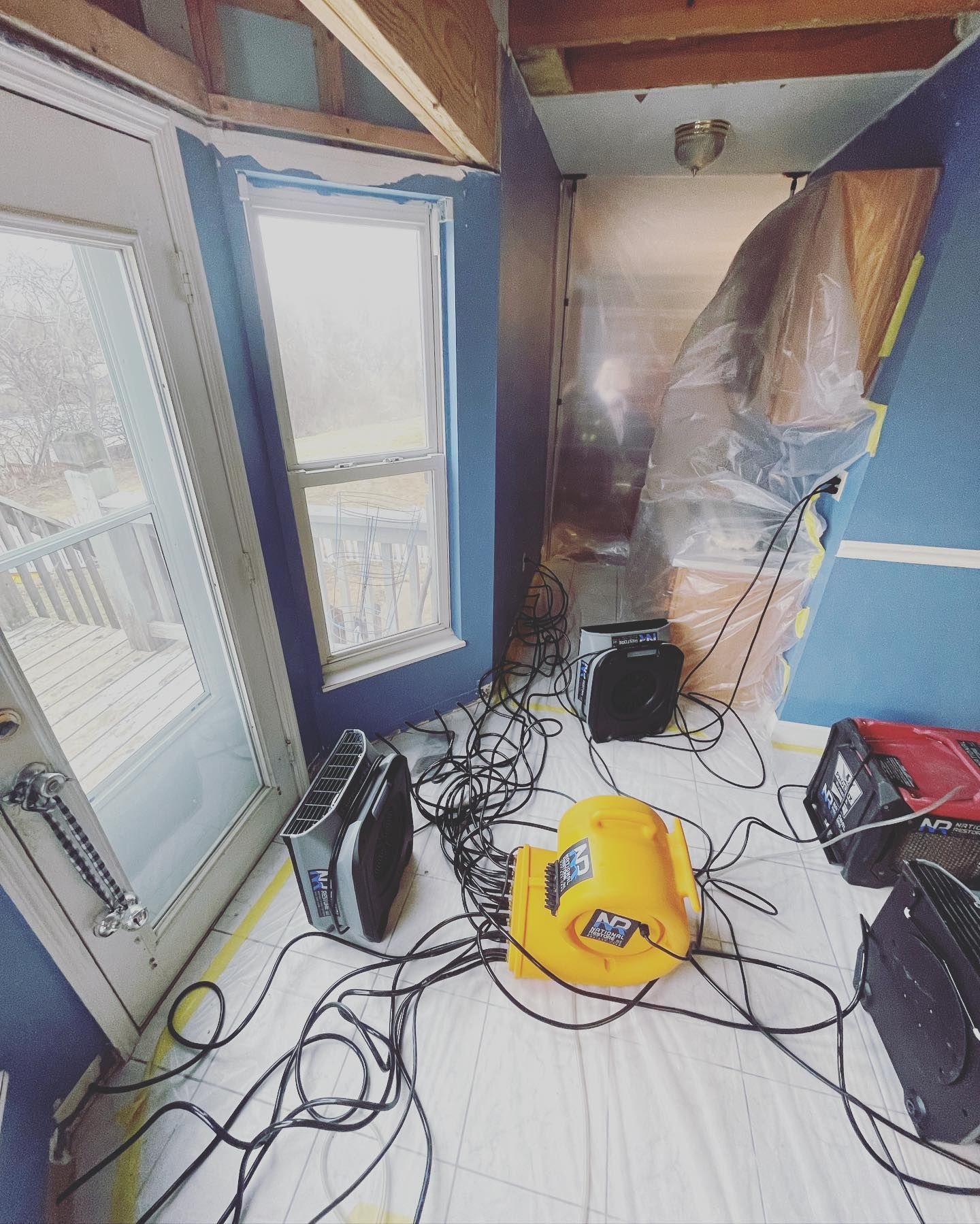 Room with blue walls and windows, drying equipment and wires present, plastic sheeting on floor.