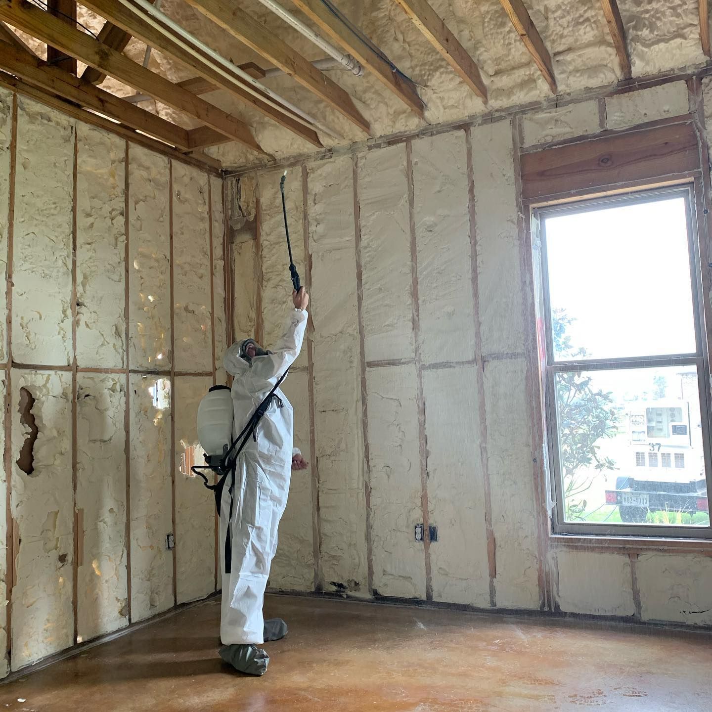 Person in protective suit spraying foam insulation in a room with a window.