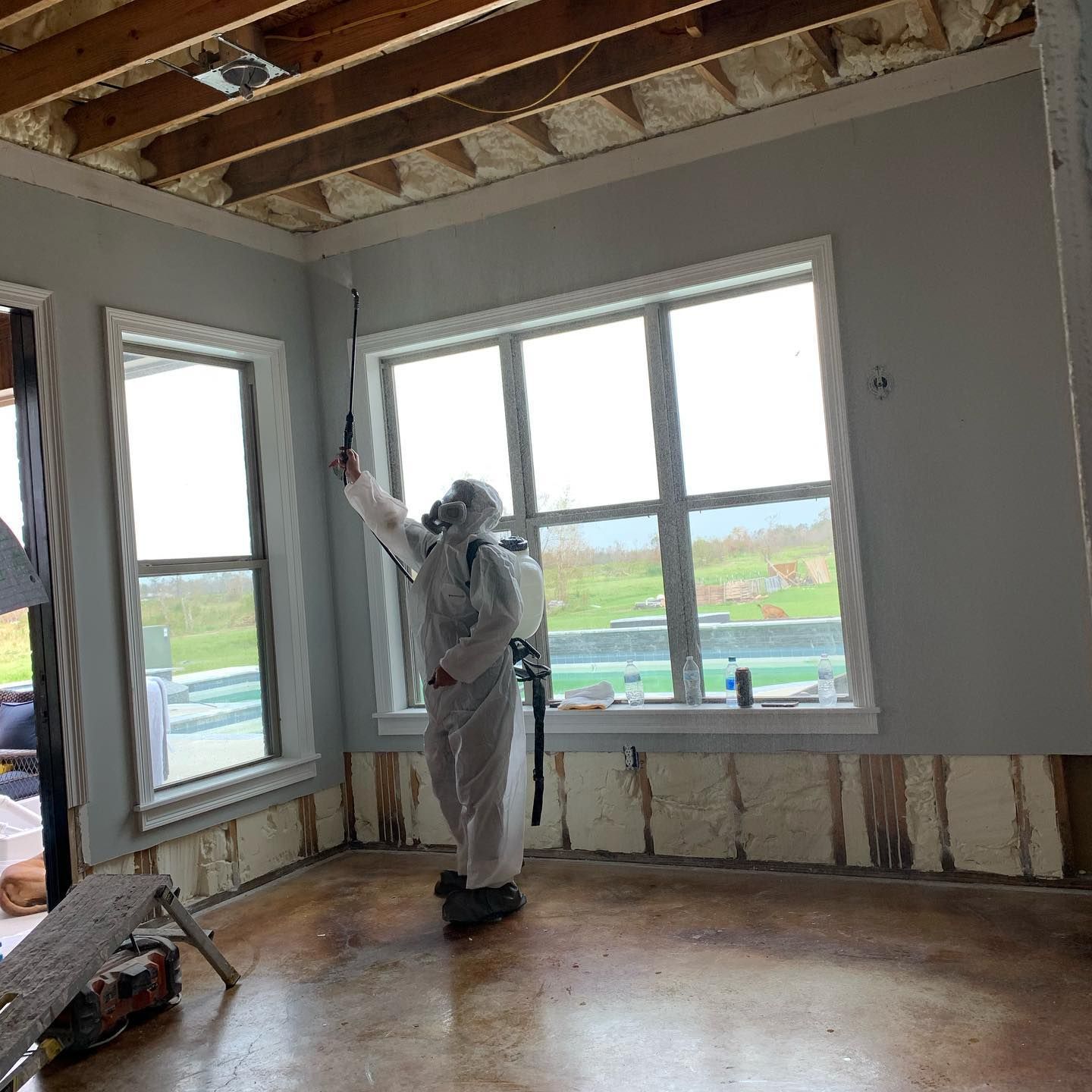 Person in protective suit spraying inside a room with exposed insulation and windows.