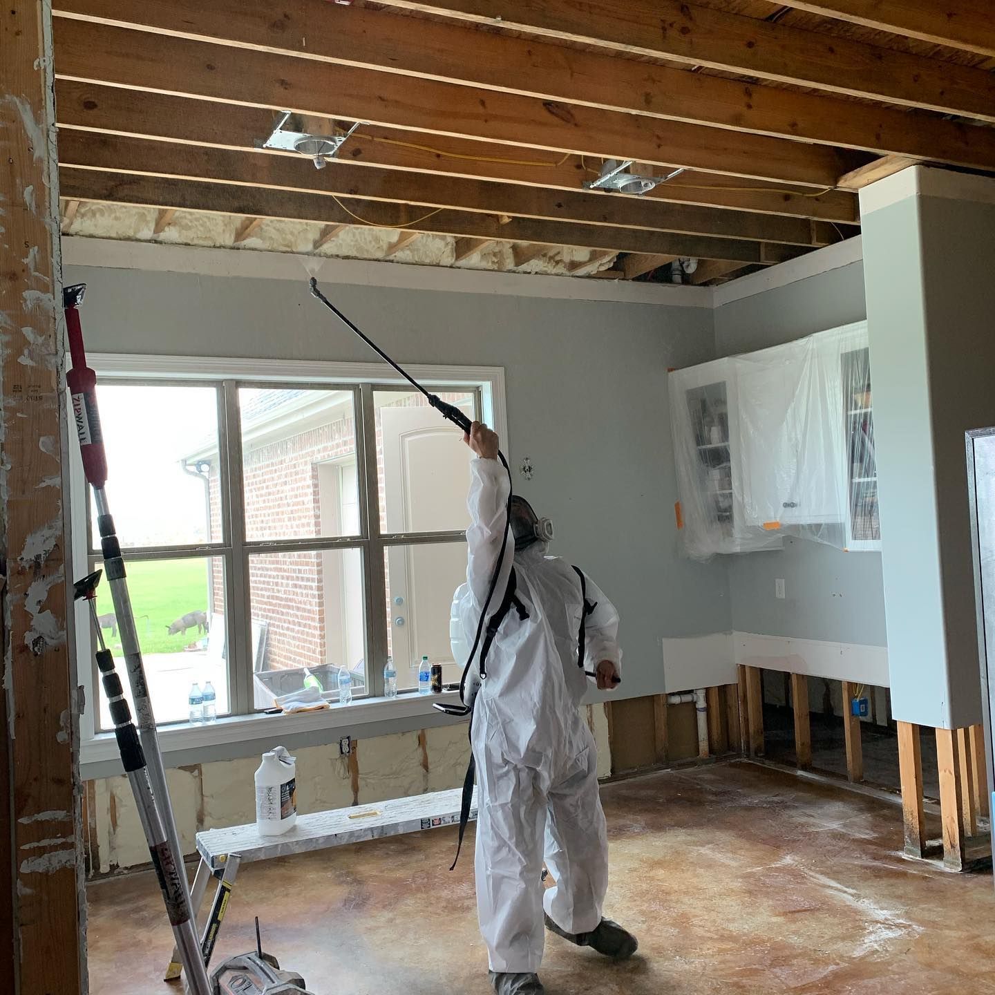 Person in protective suit spraying ceiling in a room under renovation with exposed beams and a window.