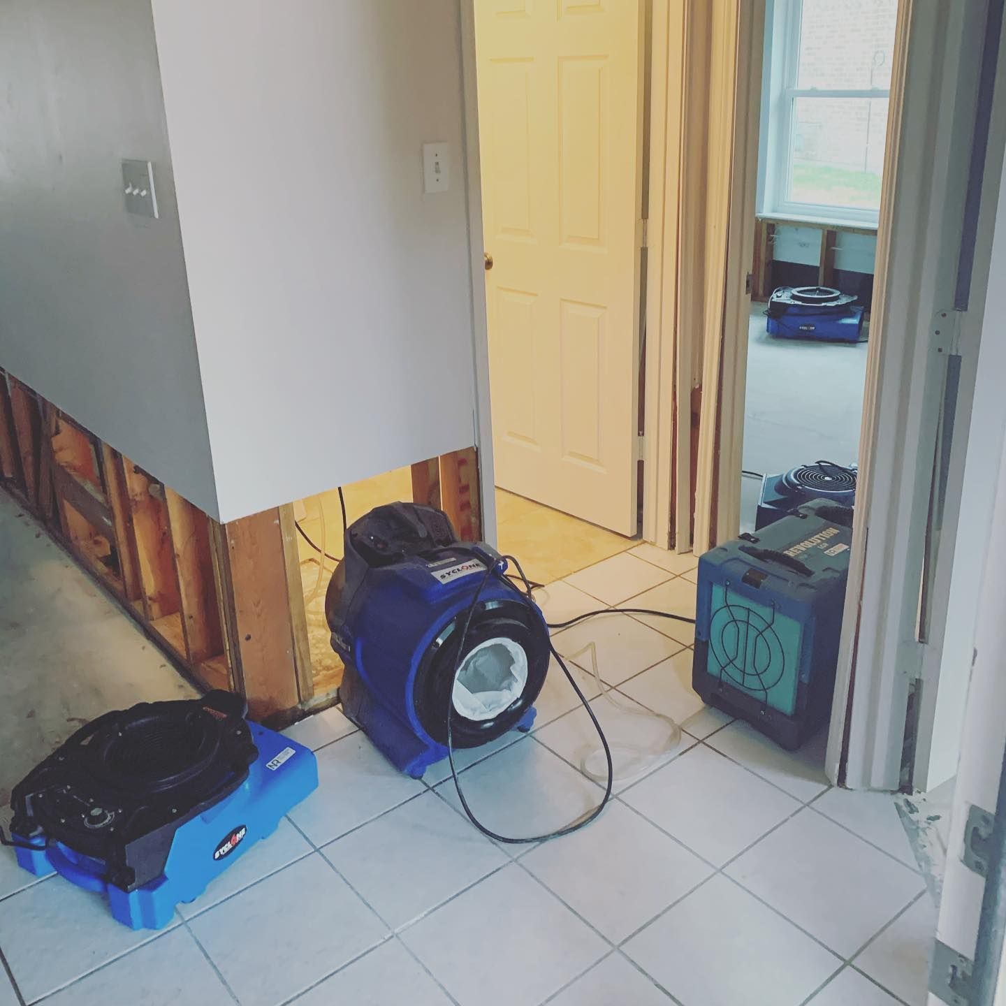 Blue fans drying a flooded hallway and room; exposed wall studs; white tile floors.