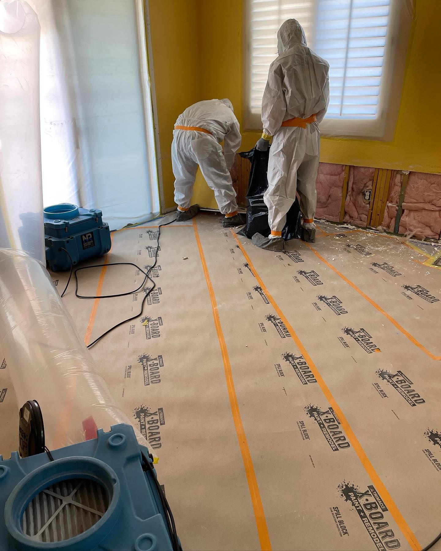 Two workers in hazmat suits remediate a flooded room. They use equipment on a plastic-covered floor.
