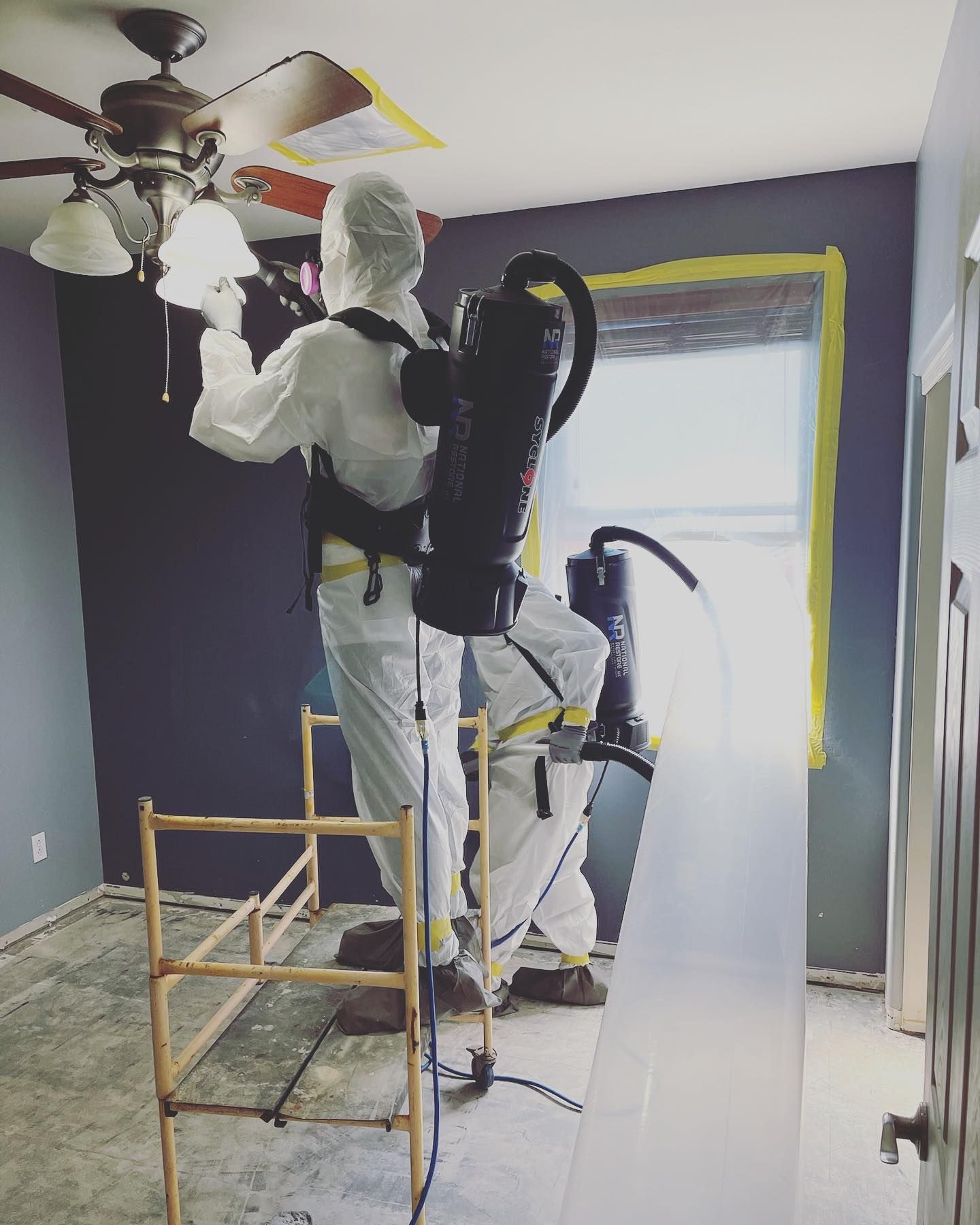 Two people in protective suits cleaning a room, one on a ladder, with a large vacuum.